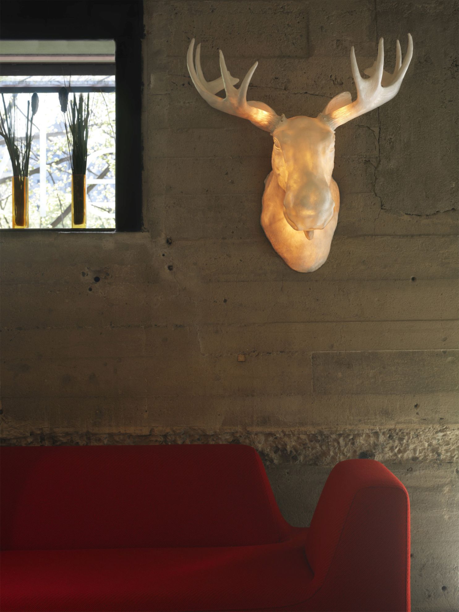 Moo Wandleuchte Northern, Elchkopf-Design, an Betonwand über rotem Sofa. Design Lampe für Wohnzimmer.