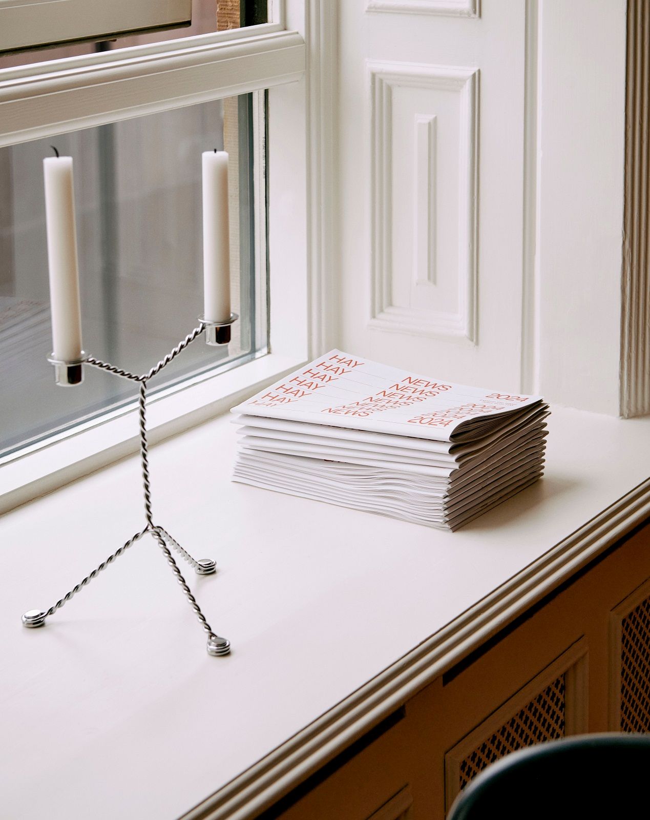 Hay Wire Candleholder Kerzenständer aus Metall mit zwei weißen Kerzen auf einer Fensterbank neben Papierstapel.