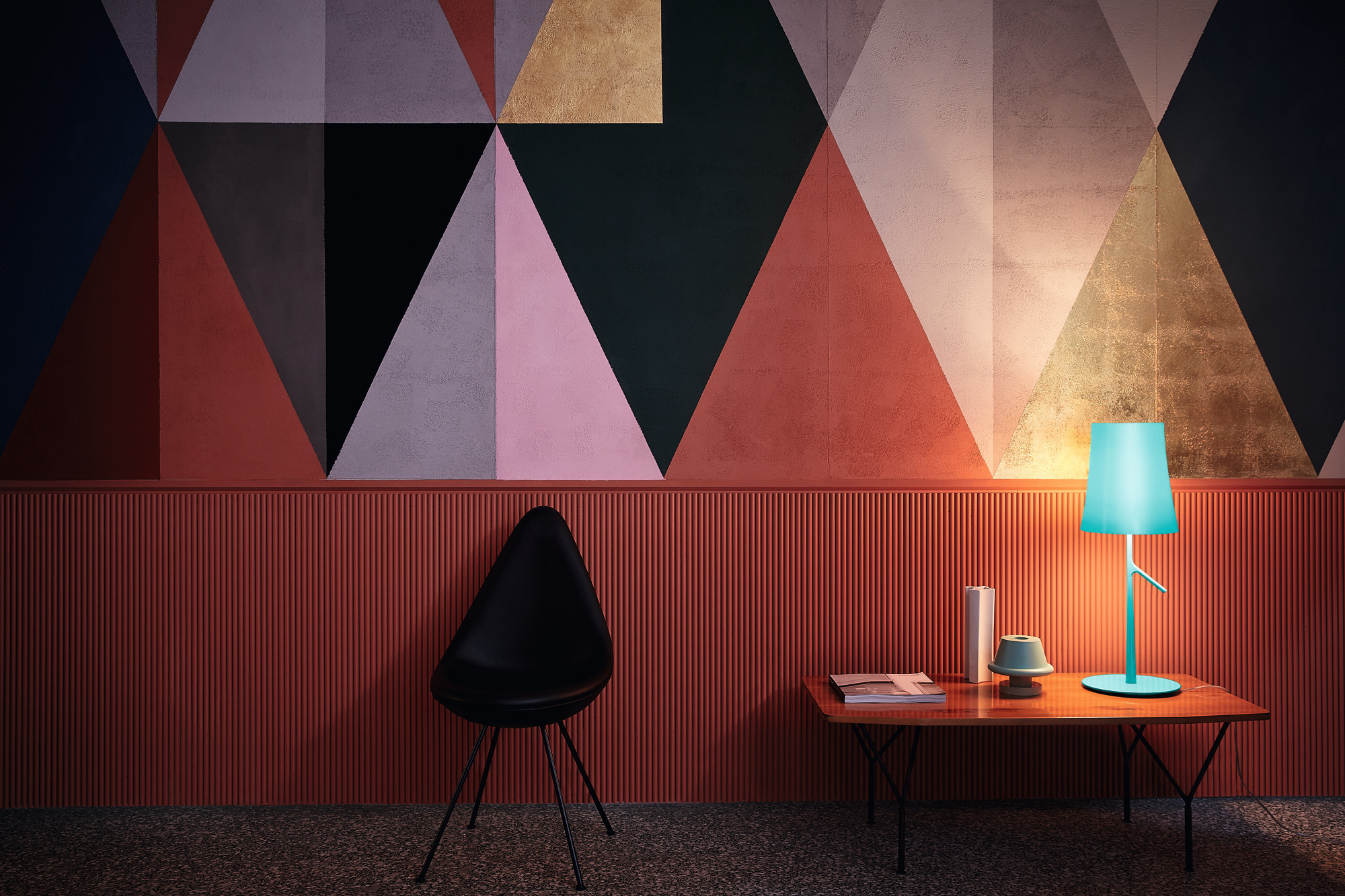 Türkisblaue Birdie Tischleuchte von Foscarini auf Holztisch vor geometrischer Wand. Design Lampe für Wohnraum.