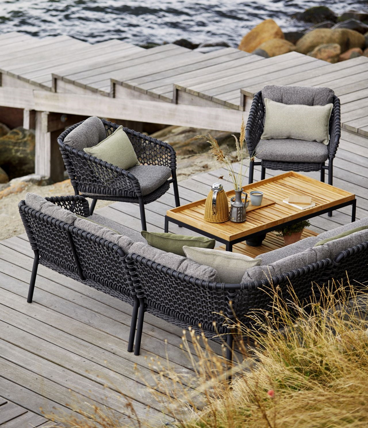 Geflochtene Gartenmöbel-Loungegruppe mit Sofa, Sesseln und Holztisch auf Holzterrasse am Meer. Outdoor-Möbel für Terrasse und Garten.