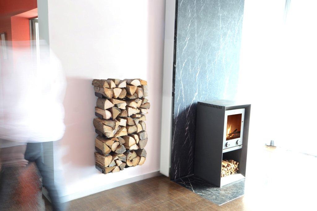 Wooden Tree Kaminholzregal Radius an weißer Wand, daneben Kaminofen mit Holz, modernes Wohnambiente.