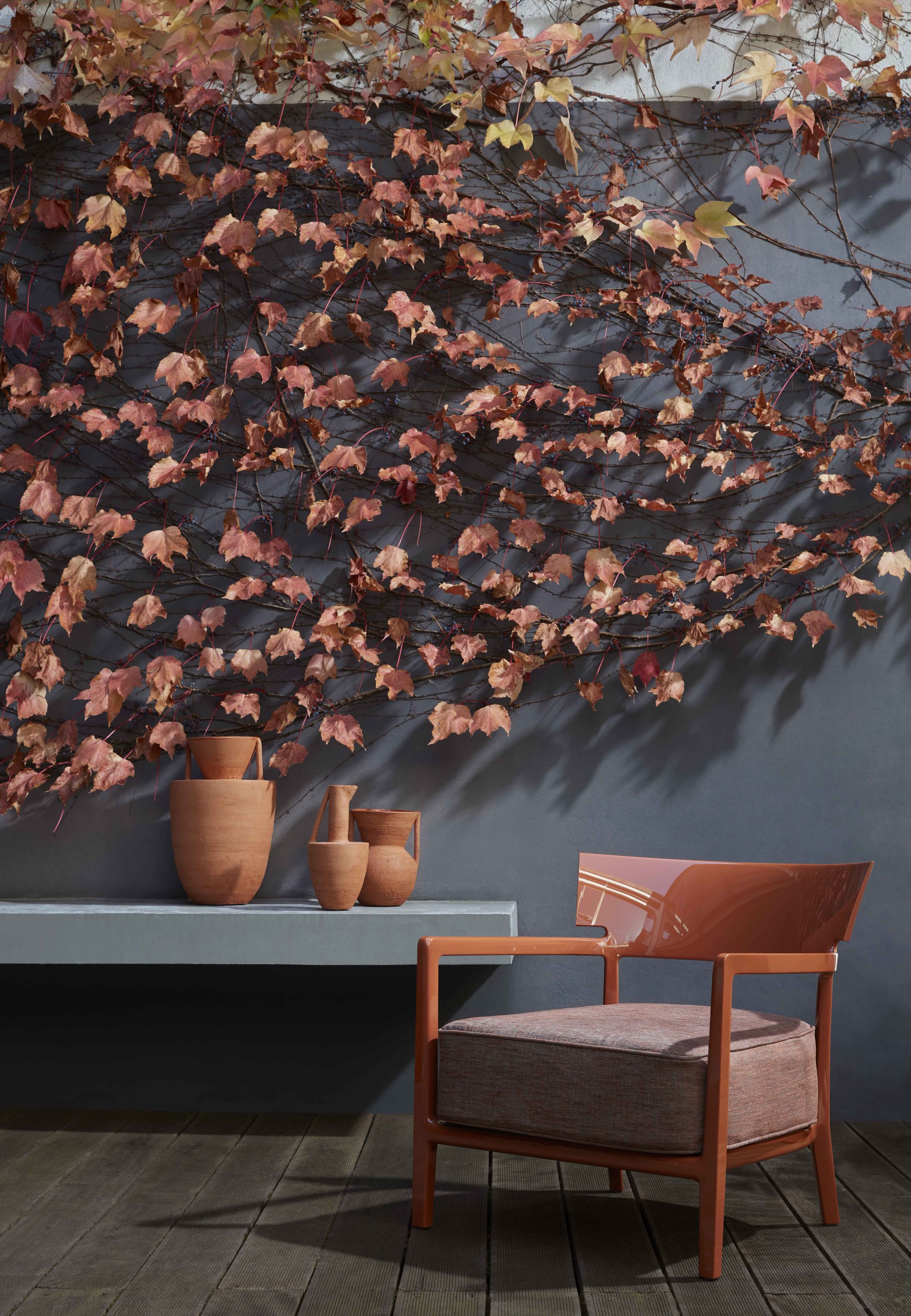 Cara Outdoor Sessel von Kartell in Terrakotta vor grauer Wand mit Efeu und Vasen.