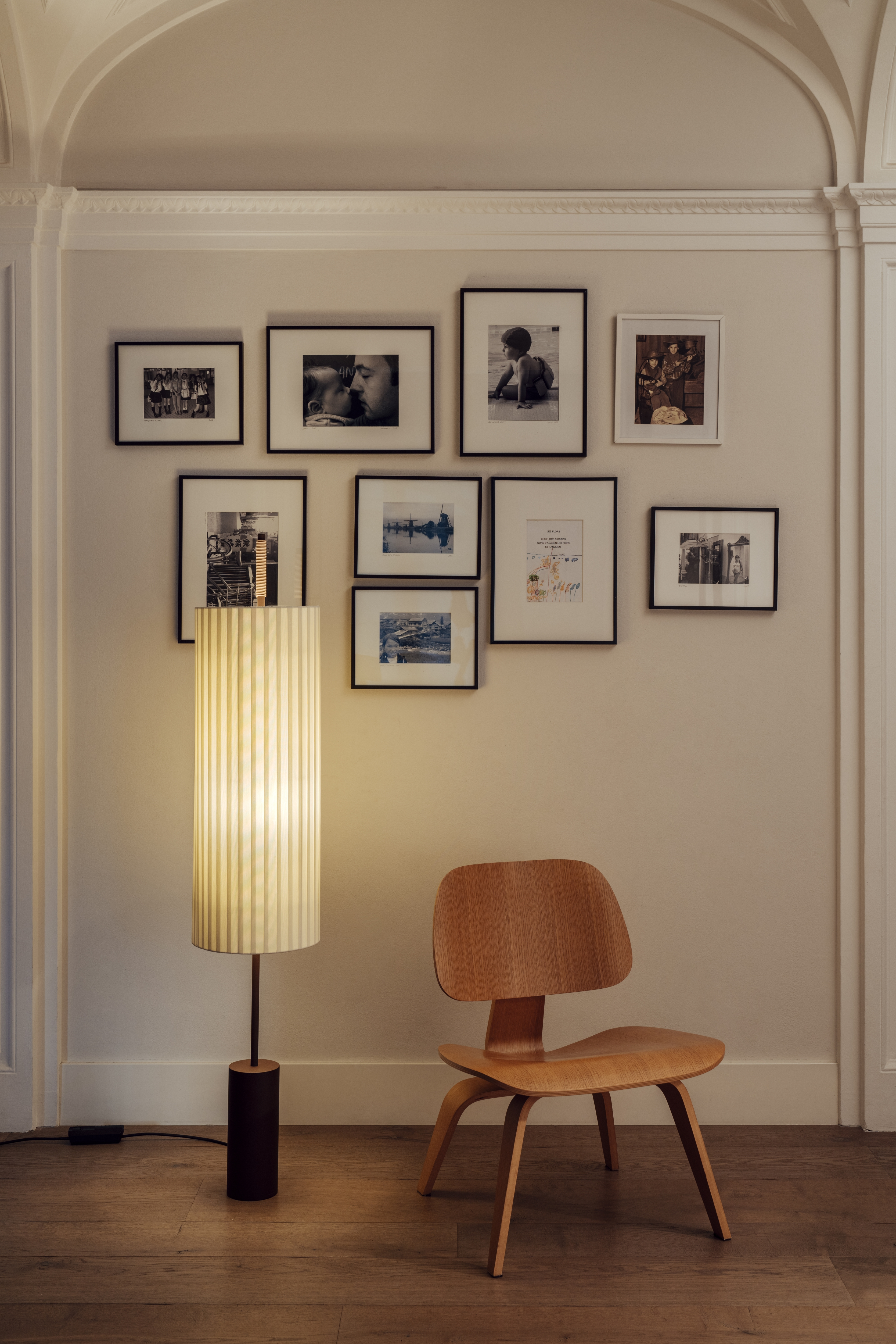 Dorica Stehleuchte von Santa & Cole neben einem Holzstuhl vor einer Bilderwand.