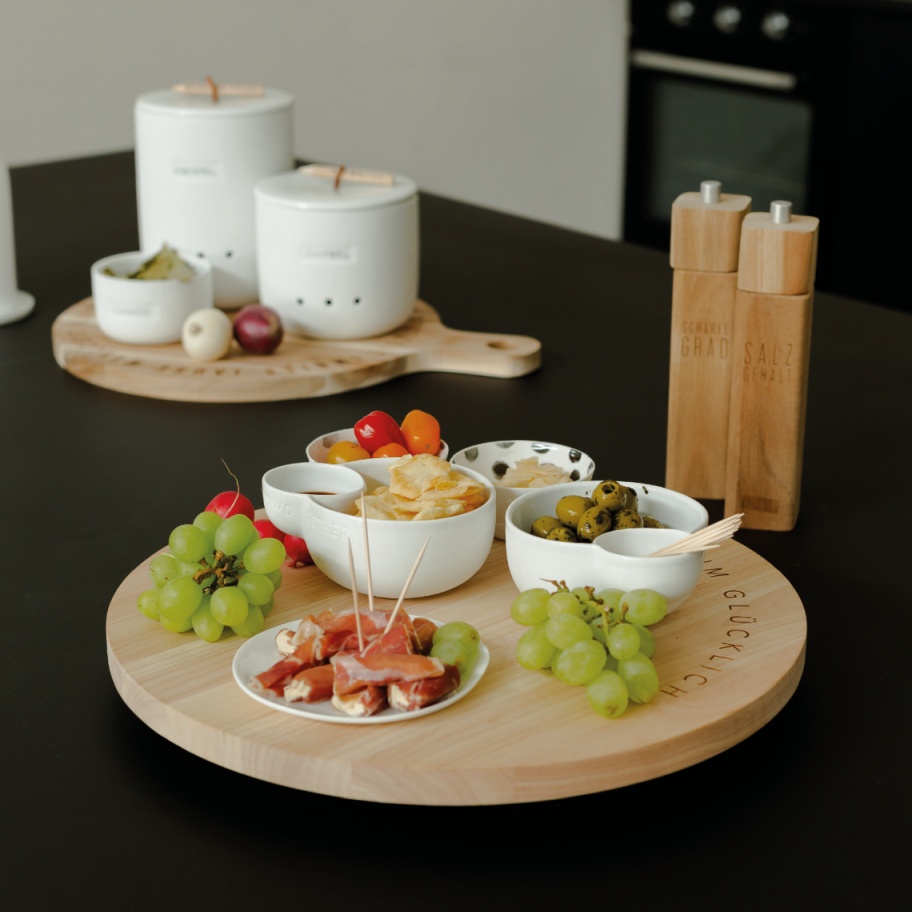 Drehplatte "Rundum Glücklich" mit Snacks: Oliven, Käse, Weintrauben und Dips in weißen Schalen. Servierplatte aus Holz.