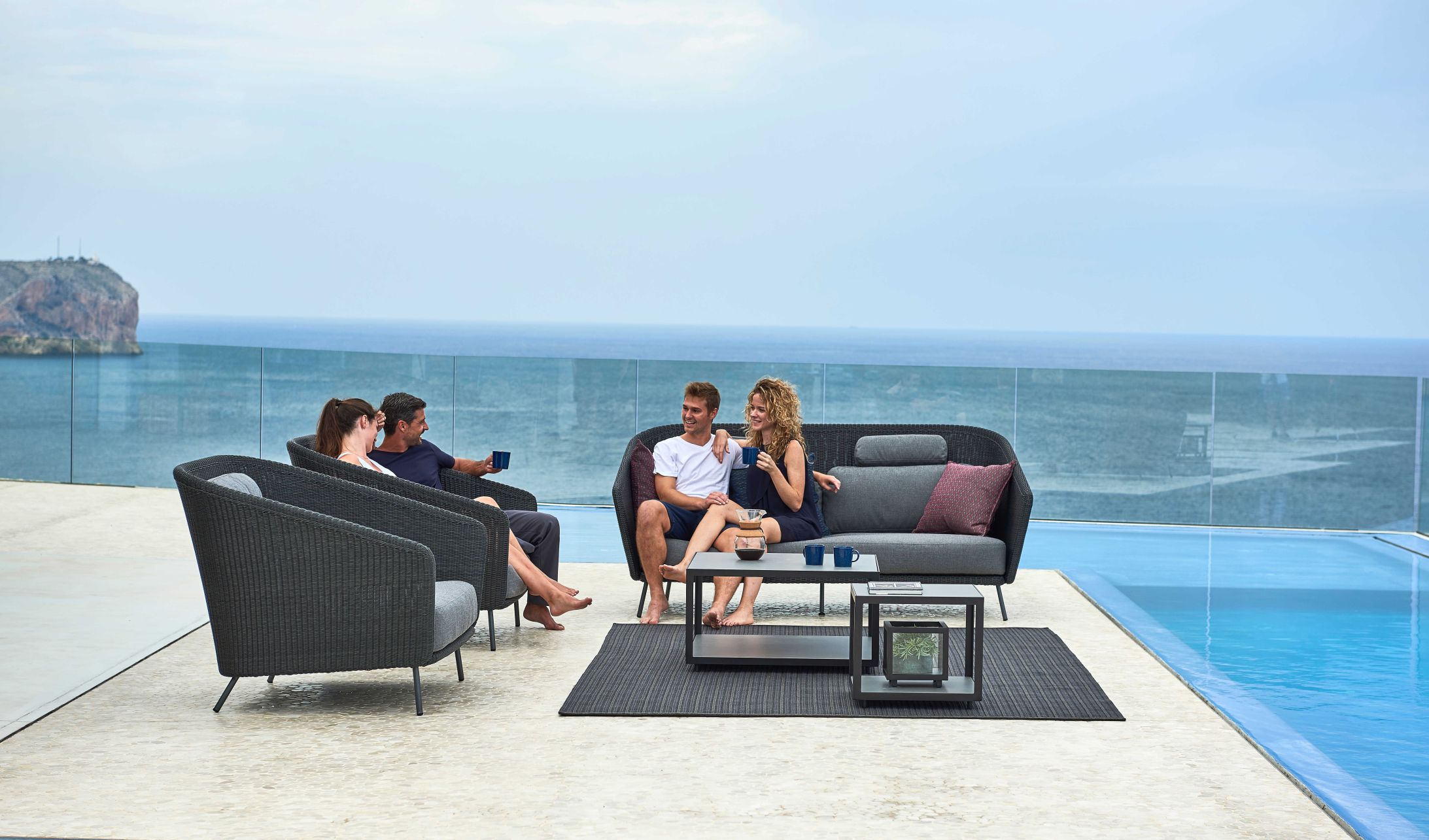 Personen entspannen auf Cane-Line Gartenmöbeln: Loungesessel, Sofa und Tisch am Pool mit Meerblick.