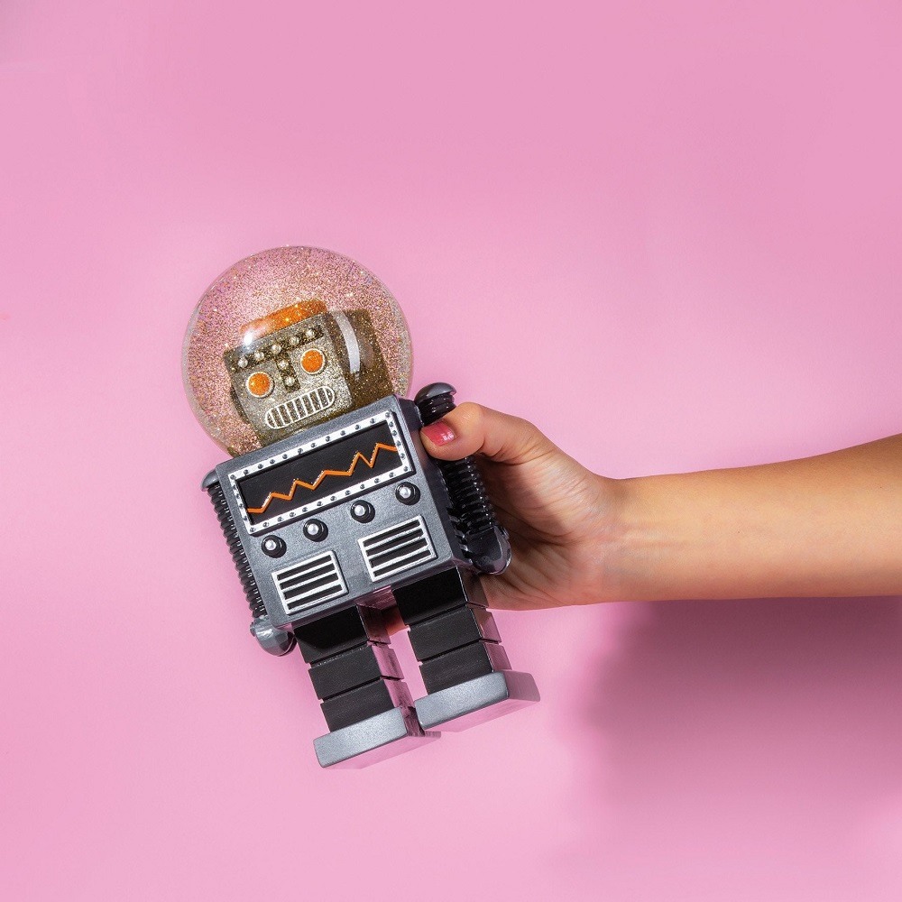 Nahaufnahme einer Hand, die eine Summerglobe Glitzerkugel mit Roboter-Motiv vor pinkfarbenem Hintergrund hält.