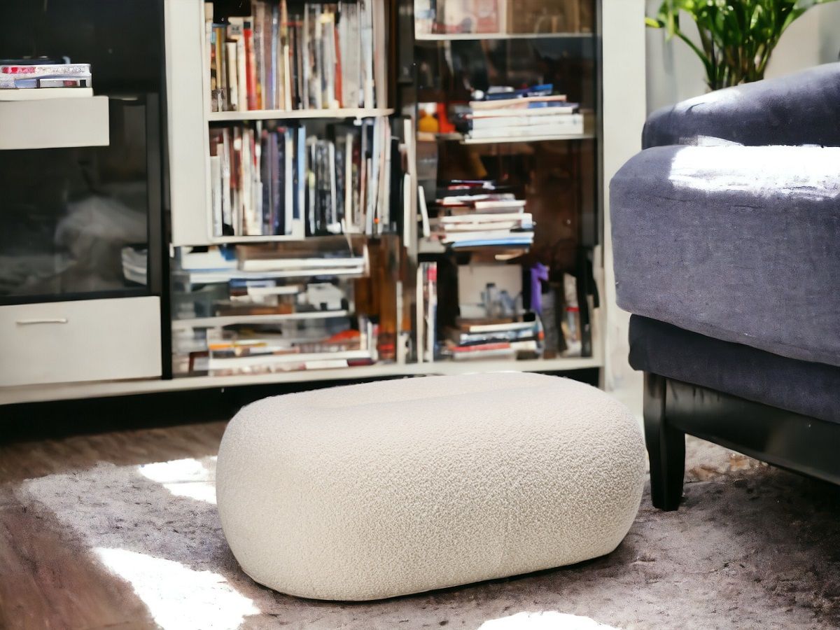 Nana Pouf Hocker von Freifrau Manufaktur, cremefarben, im Wohnzimmer auf Teppich vor Bücherregal.