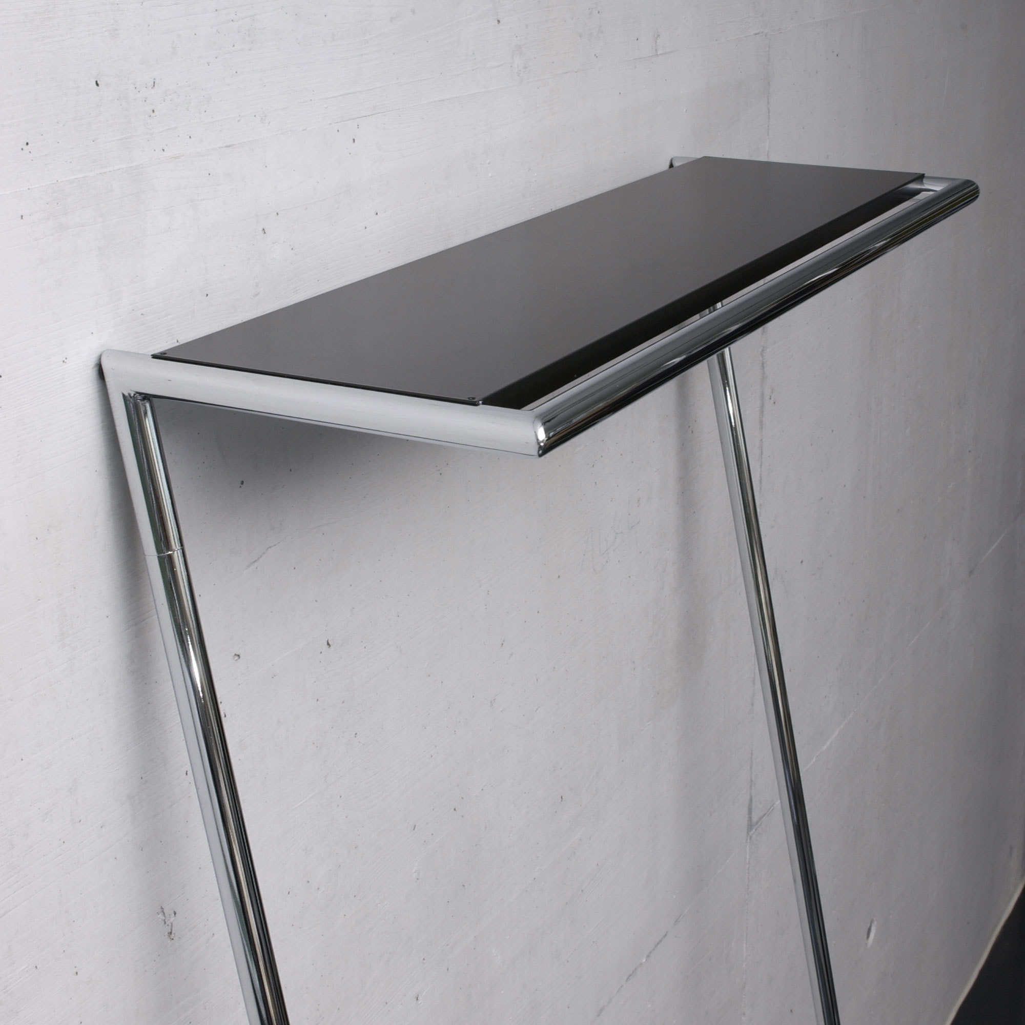 Schwarze Hutablage für Zen und Link Garderobe von Mox mit verchromtem Gestell vor einer Betonwand.