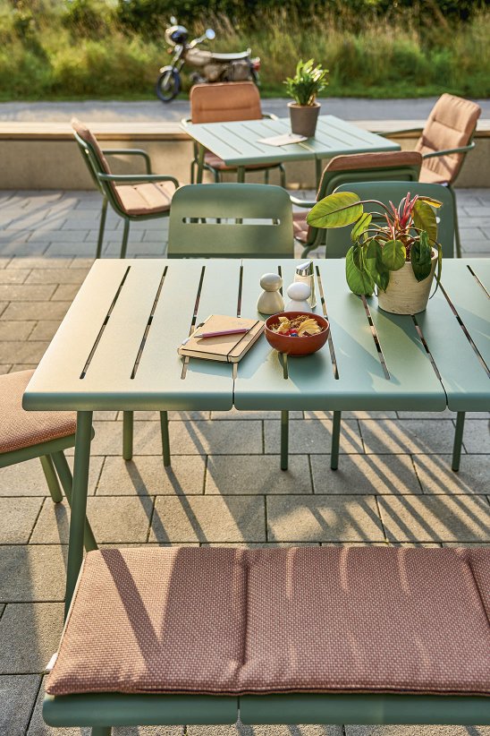 Weishäupl Vapio Stool Hocker Outdoor: Gartentisch mit Stühlen und Bank in Grün, gedeckt mit Schüssel, Notizbuch und Pflanze.