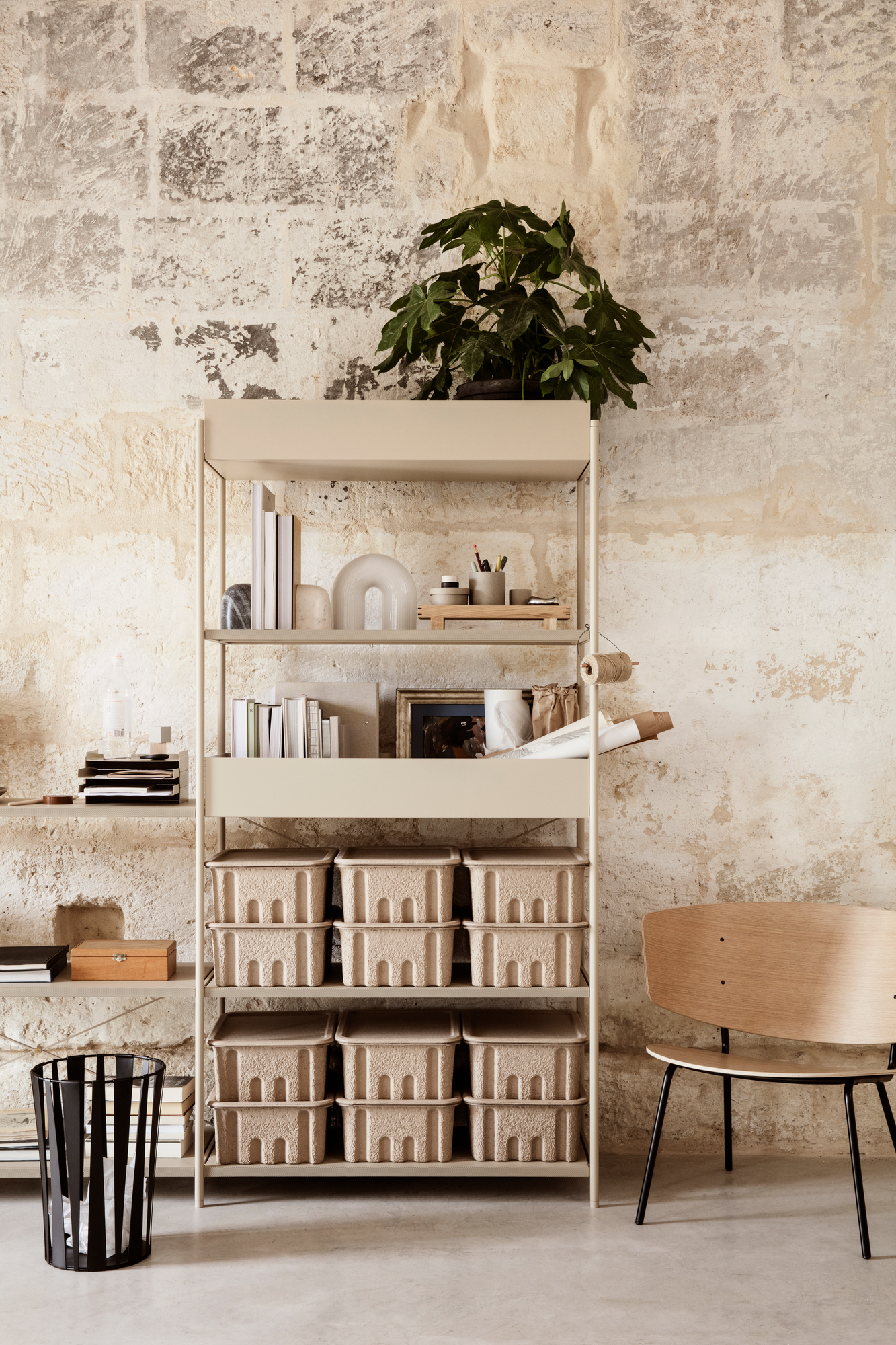 Beige Ferm Living Regalsystem mit Aufbewahrungsboxen, Dekoration und Pflanze vor einer strukturierten Wand.