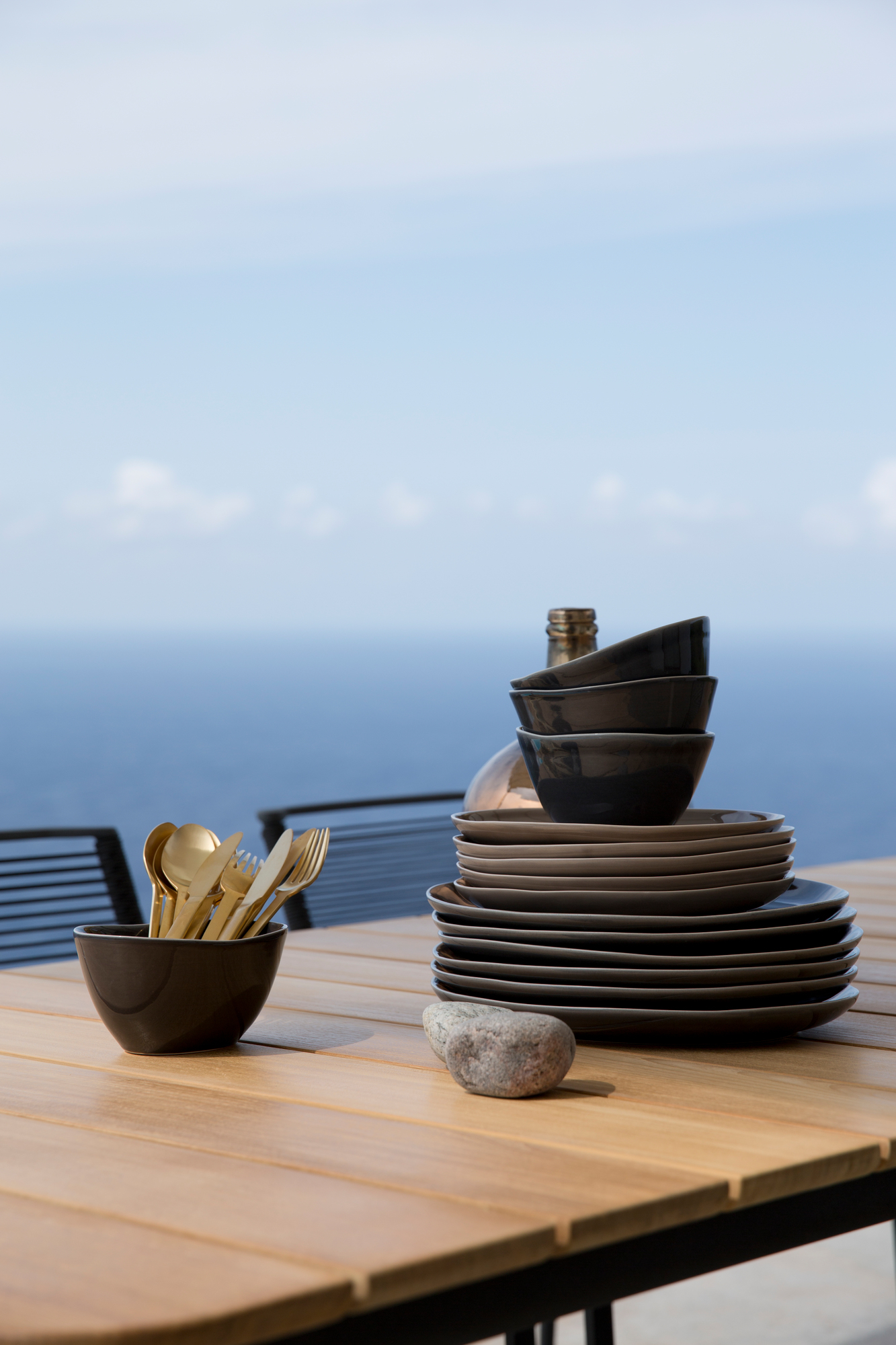 Cane-Line Core Outdoor Esstisch Teak mit Geschirr und goldenem Besteck vor Meereskulisse.