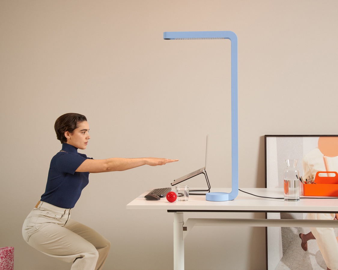 Frau macht Dehnübungen am Schreibtisch unter der grauen Team Home Tischleuchte für ein ergonomisches Büro.