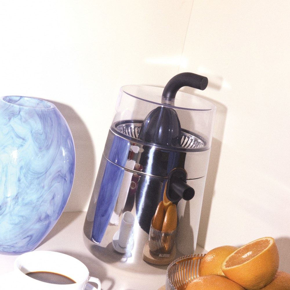 Alessi NE02 B Toru elektrische Zitronenpresse aus Edelstahl mit Orangen und Tasse Kaffee.