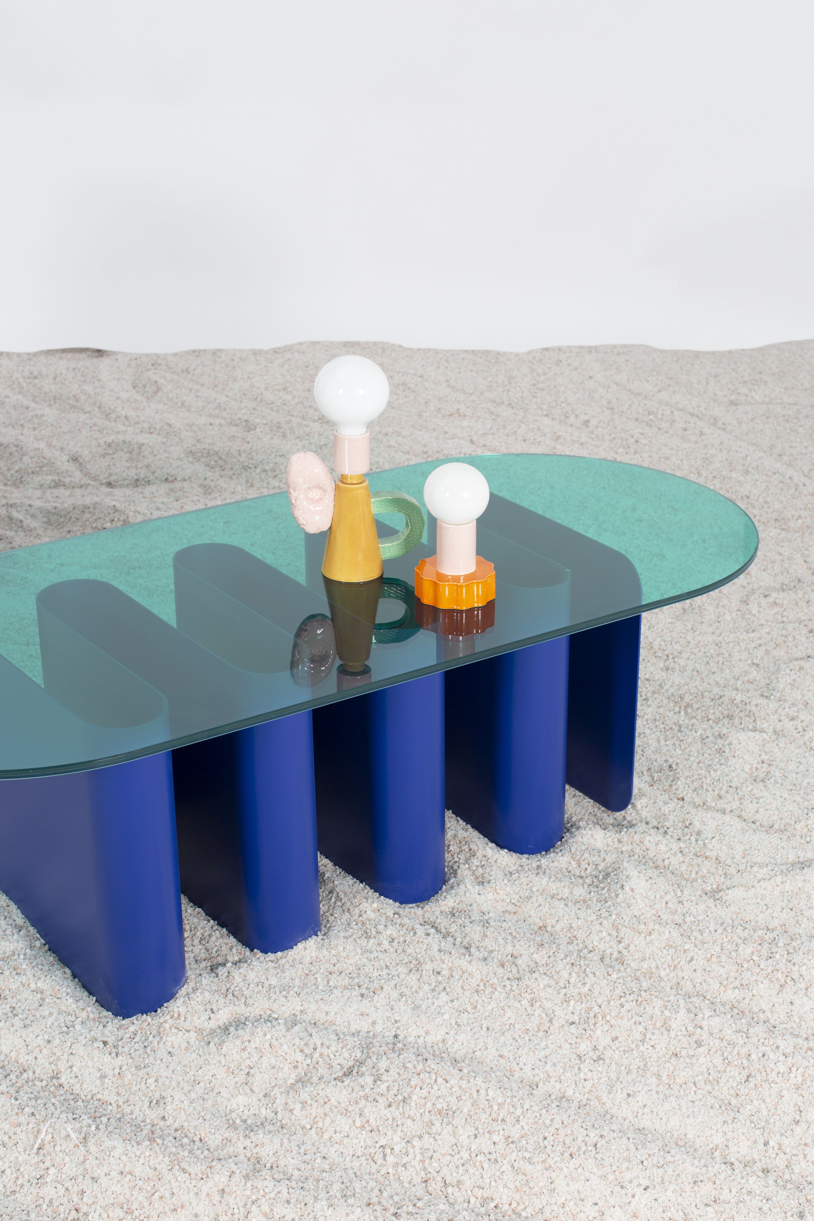 Tavolo2 Esstisch Pulpo mit blauen Beinen und Glasplatte, darauf zwei Lampen und ein Stein.