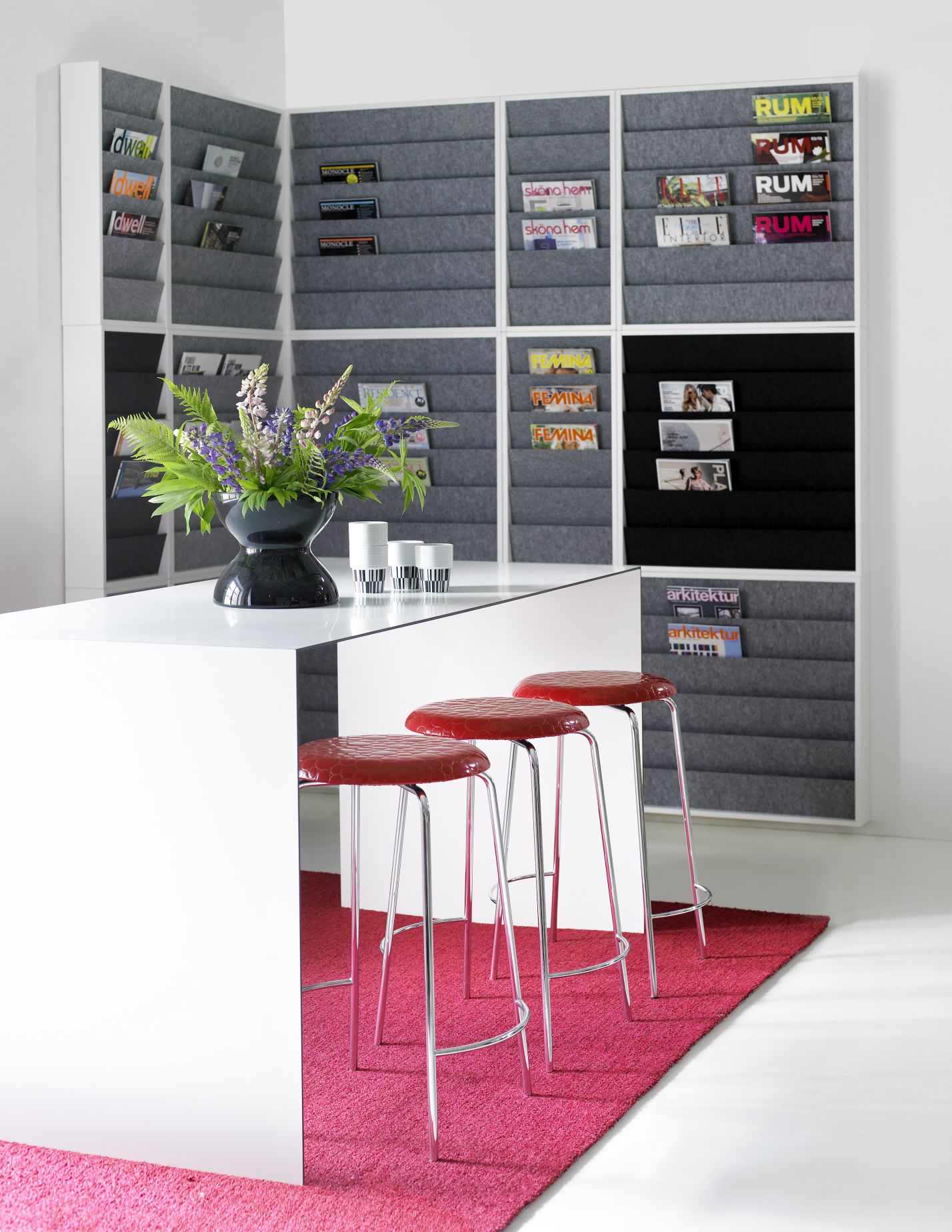 Modernes Büro Interieur mit Magazinwand, weißem Tresen, roten Barhockern und pinkem Teppich.