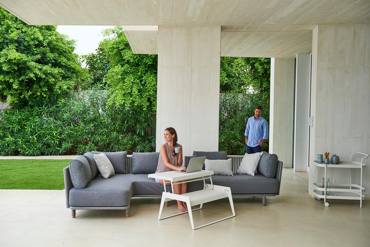 Frau nutzt Laptop auf Cane-Line Moments Outdoor Sofa. Graues 2-Sitzer Gartensofa mit Kissen.
