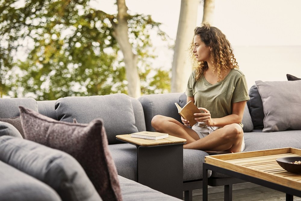 Cane-Line Beistelltisch Mega Outdoor: Frau liest auf Sofa mit kleinem Tisch.