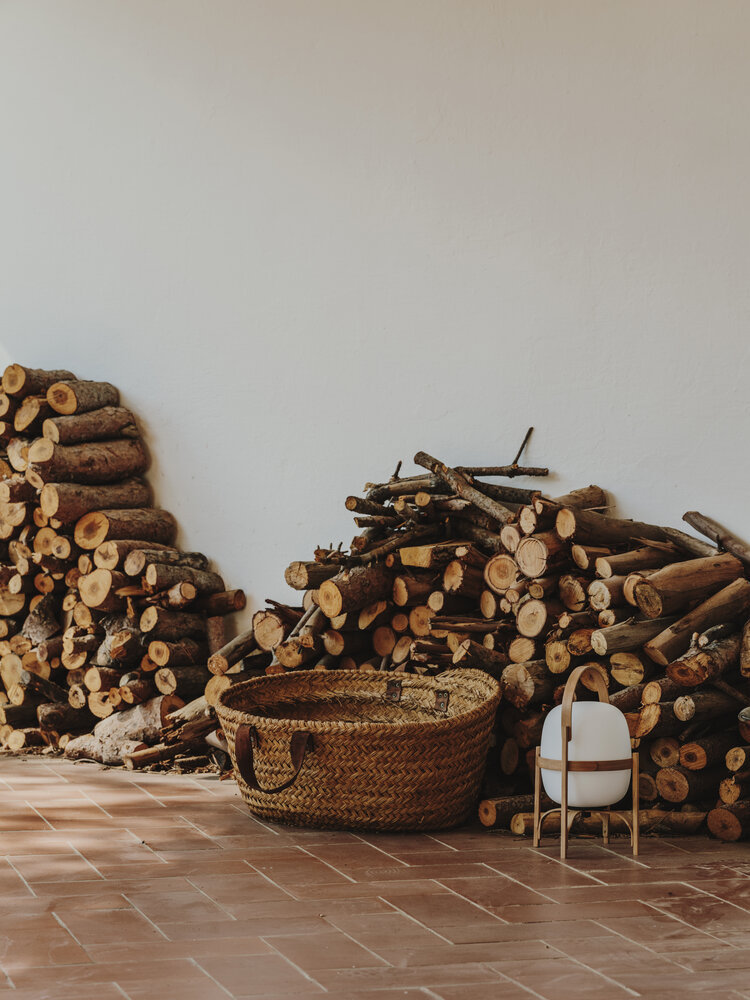 Cestita Batería Akkuleuchte von Santa & Cole neben Holzkorb und Brennholz auf Terrakottafliesen.