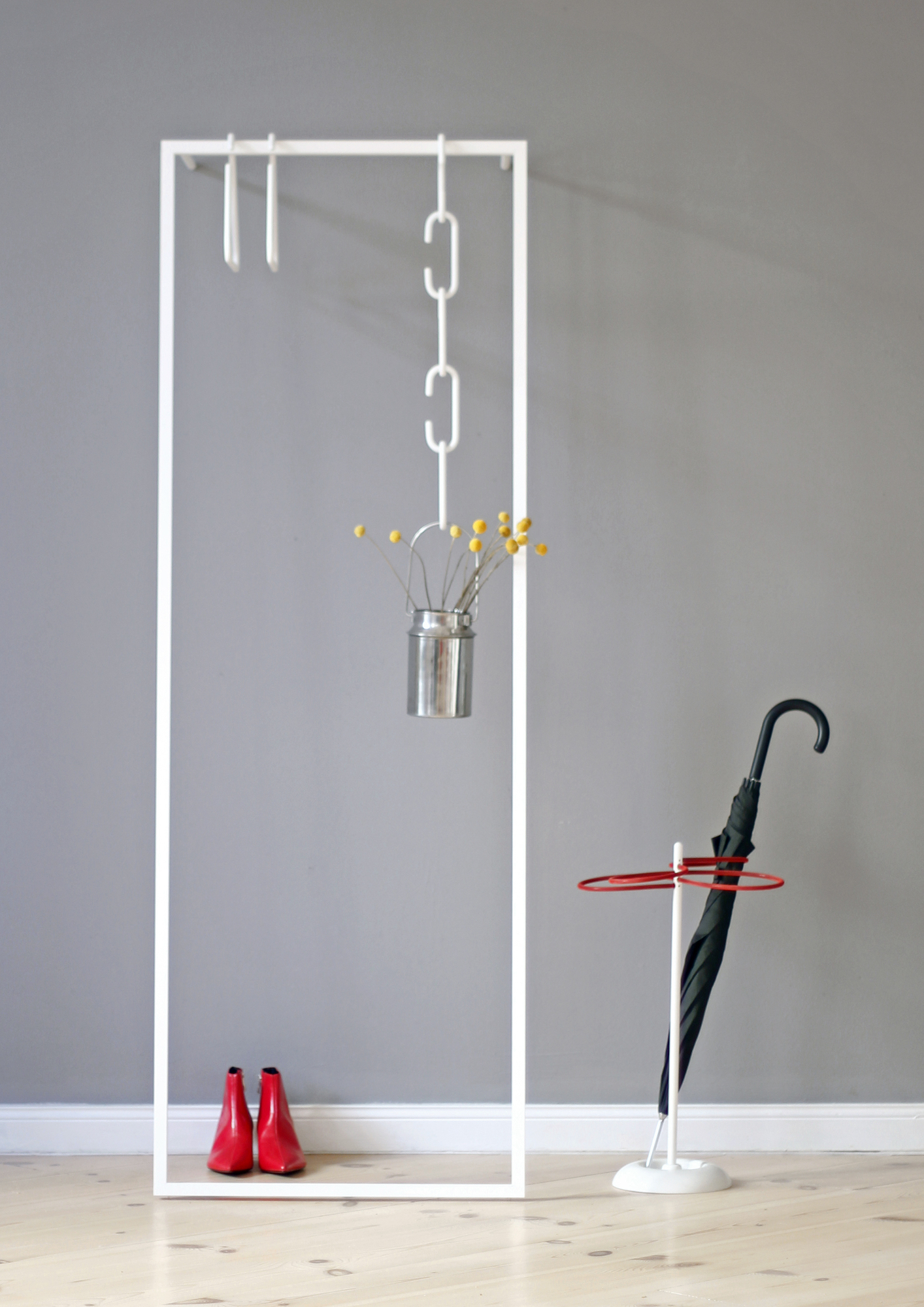 Weiße Leano Anlehngarderobe von Roomsafari mit Regenschirm, roten Schuhen und Dekoration vor grauer Wand.