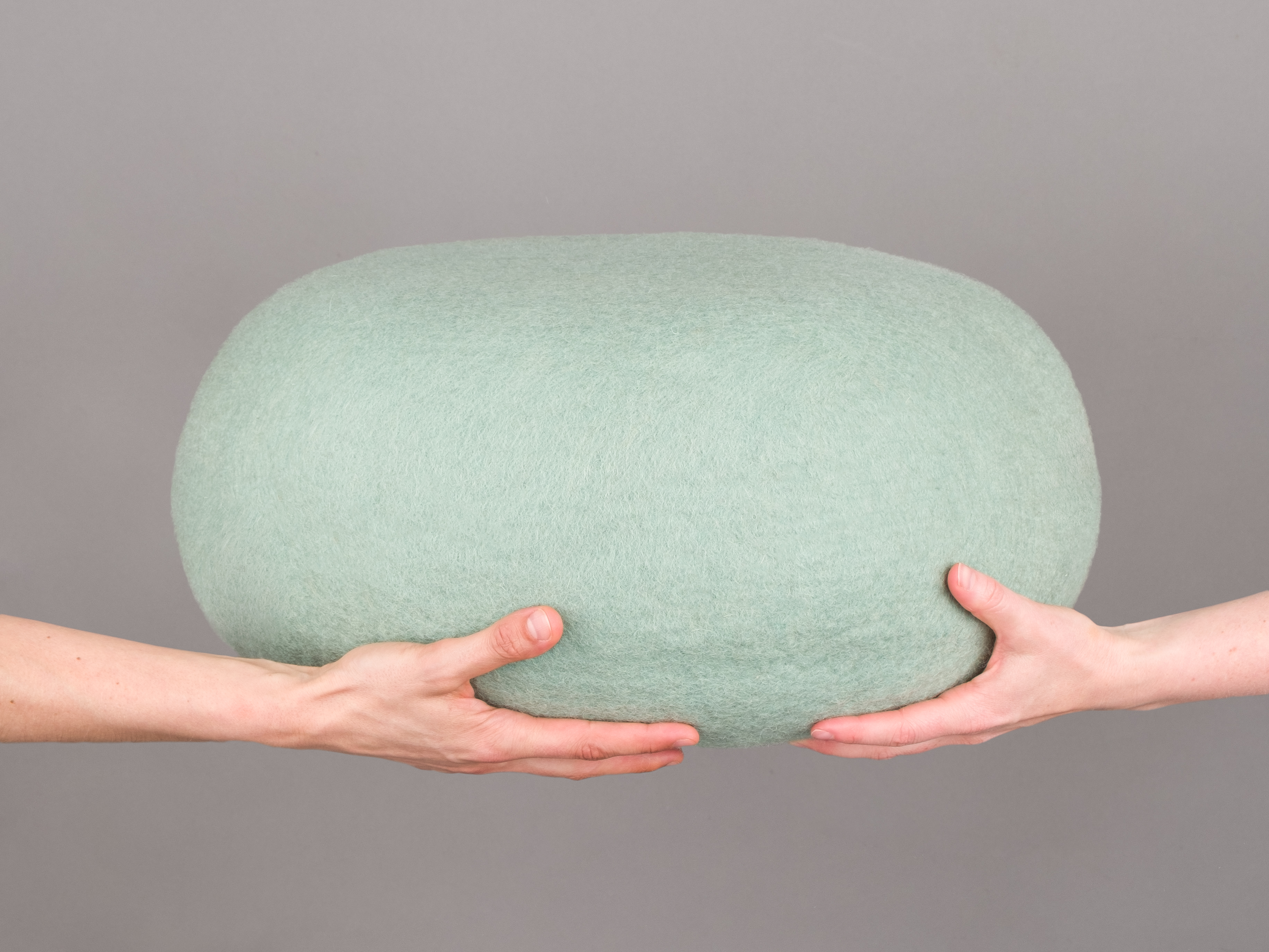 Hände halten einen runden, hellgrünen Filz Pouf Hocker. Gemütliches Wohnaccessoire für Sitzgelegenheiten.