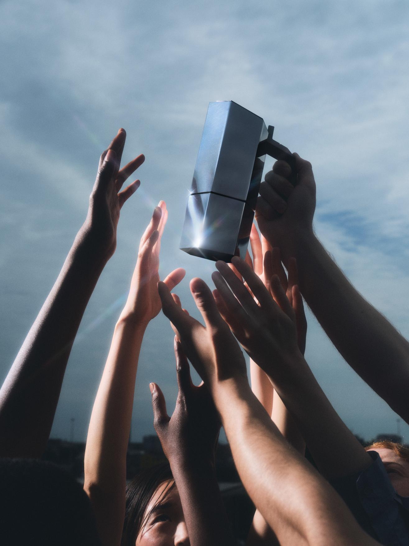 Mehrere Hände greifen nach einem silbernen Menhir Espressokocher von Alessi vor bewölktem Himmel.