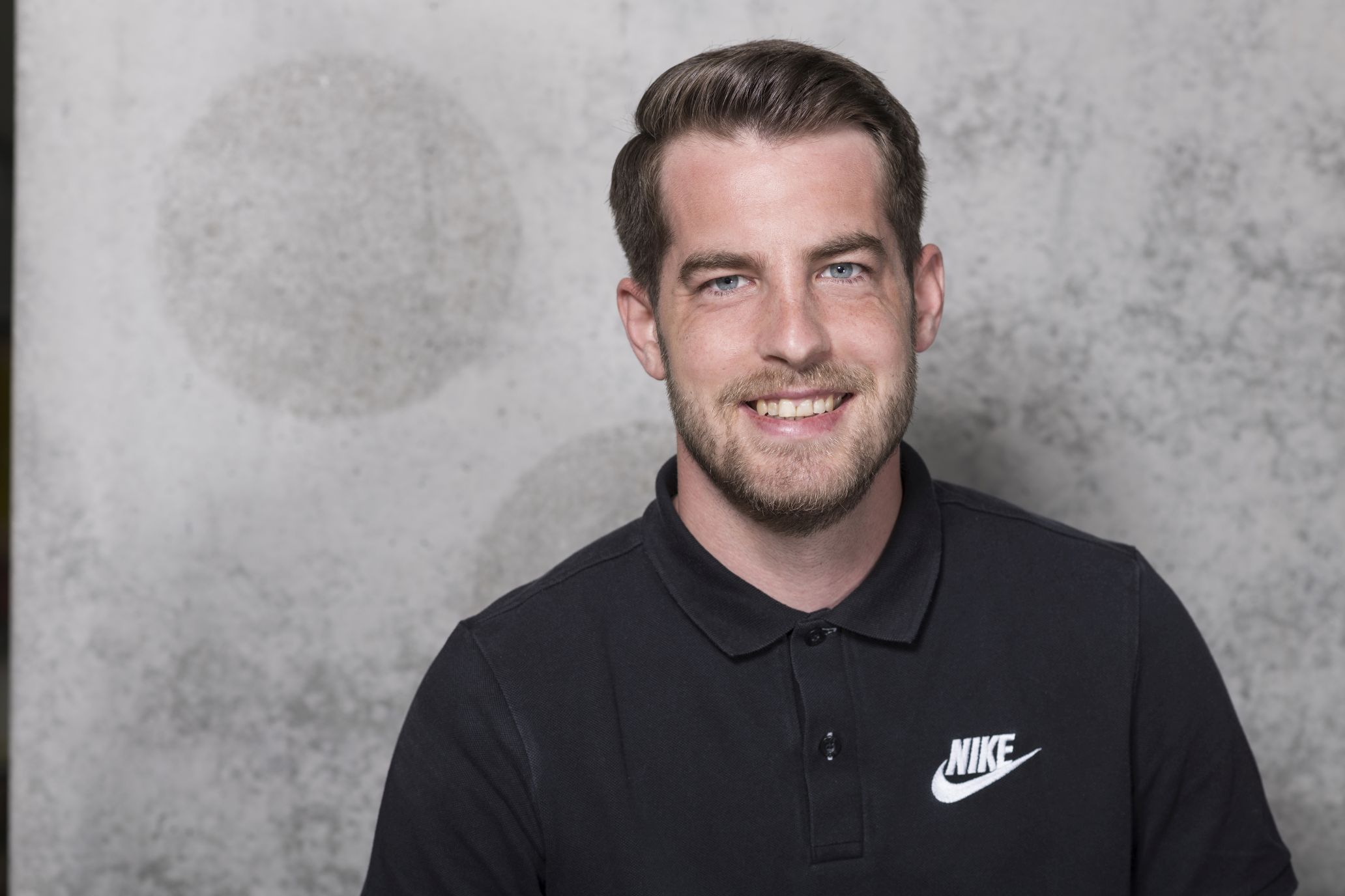 Mann mit Bart trägt schwarzes Nike Polo Shirt, lächelt in die Kamera.