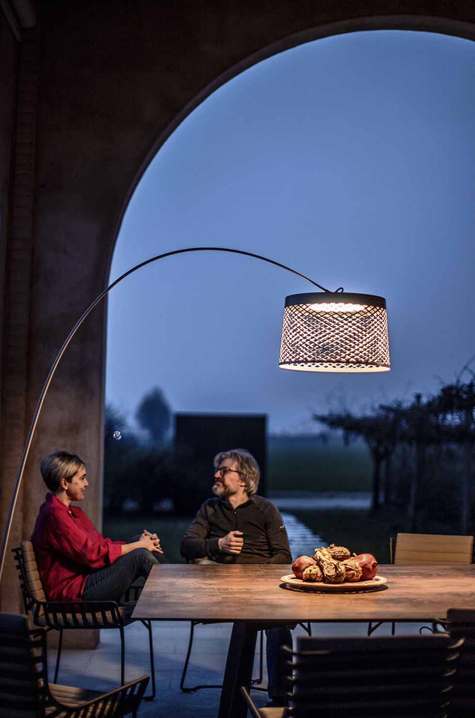 Twiggy Grid Stehleuchte kaminrot von Foscarini beleuchtet abendliche Szene mit zwei Personen und Obstschale.