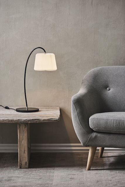 Snowdrop Tischleuchte von Le Klint mit plissiertem Schirm auf einem Holztisch neben einem Sessel.