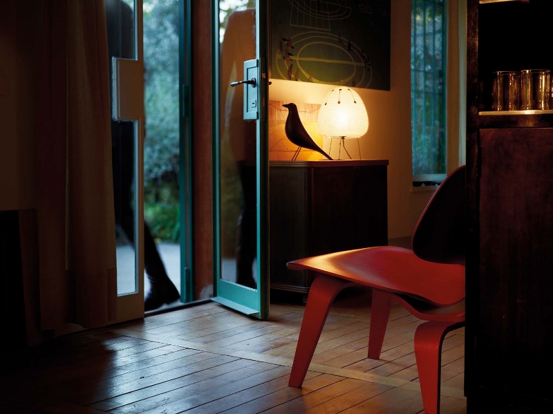 Interieur mit Eames House Bird Skulptur, Lampe und rotem Stuhl auf Holzboden. Dekorative Wohnaccessoires.