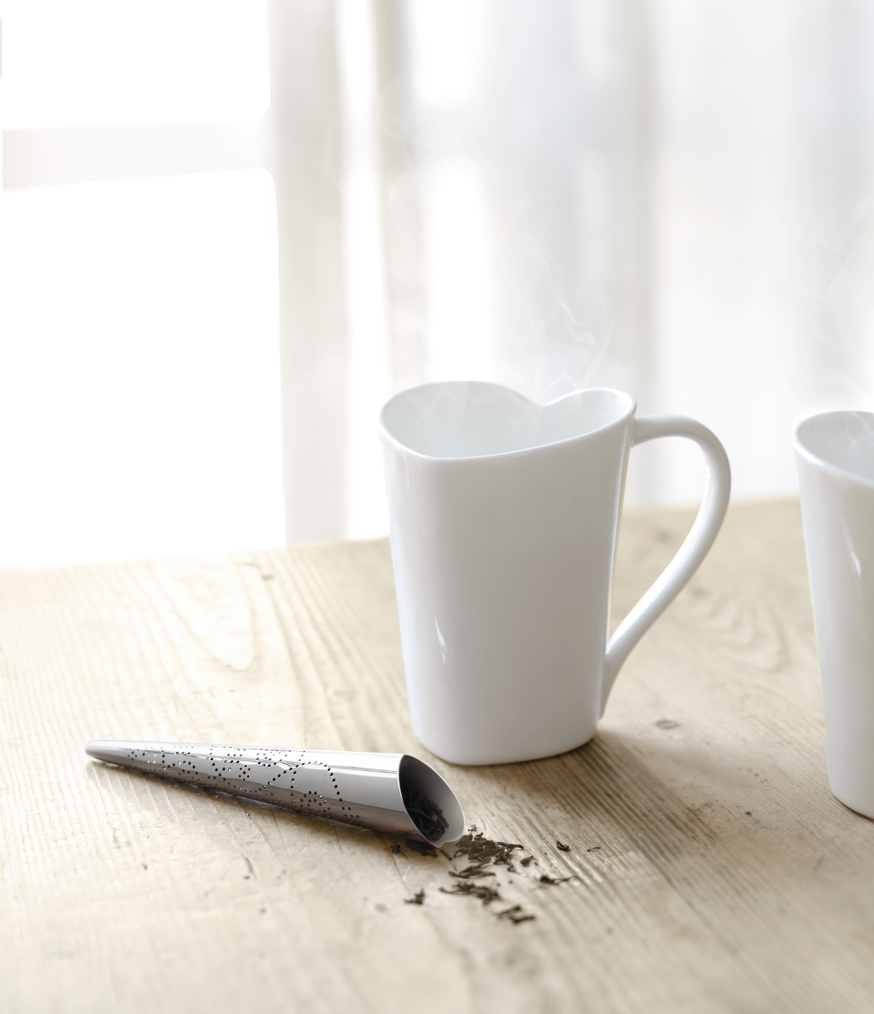 Produktfoto: Weiße Alessi Tasse mit Herzform und Teesieb auf hellem Holztisch.