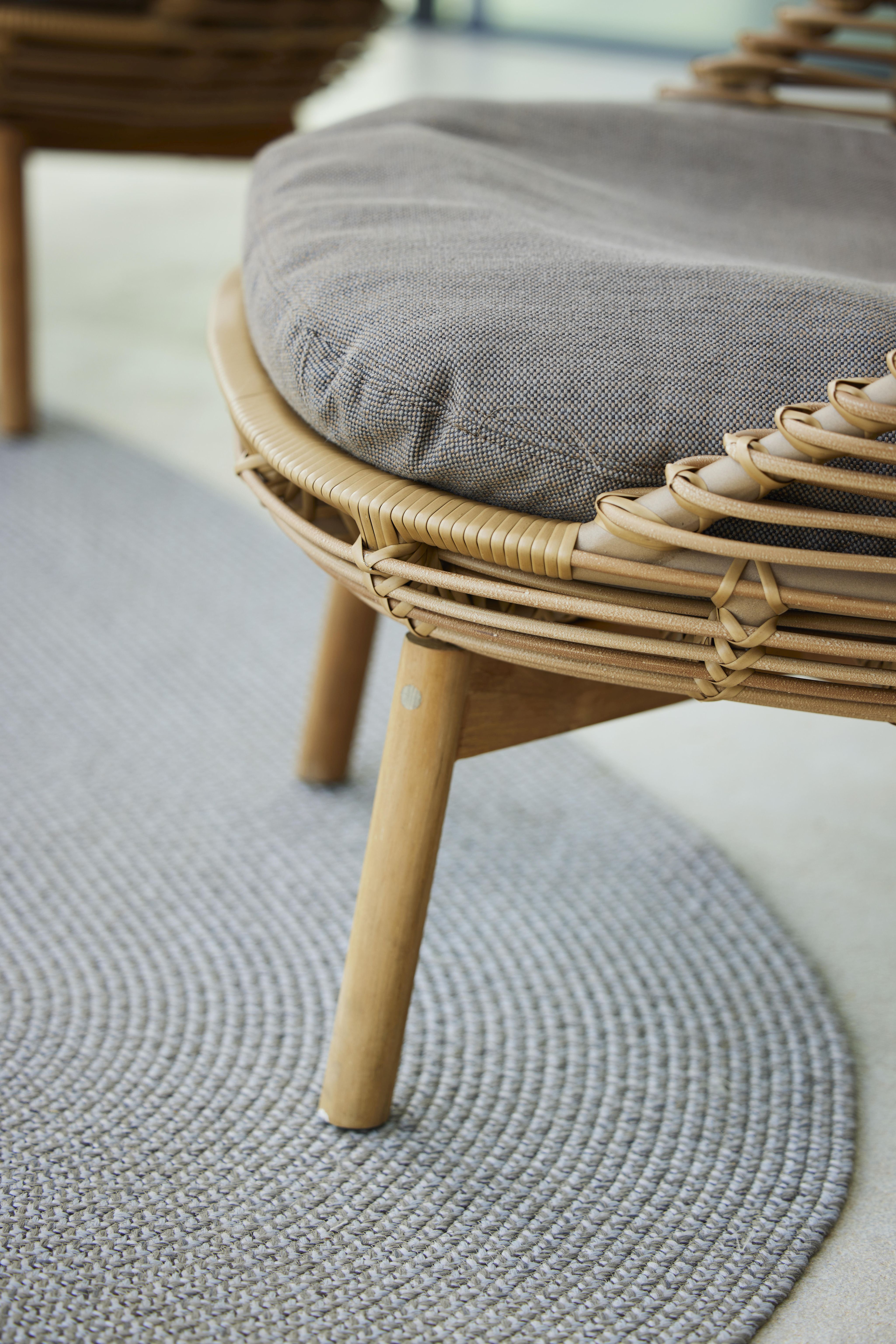Detailansicht: Cane-Line Hive Sessel mit Teakholzbeinen und grauem Kissen auf rundem Teppich.