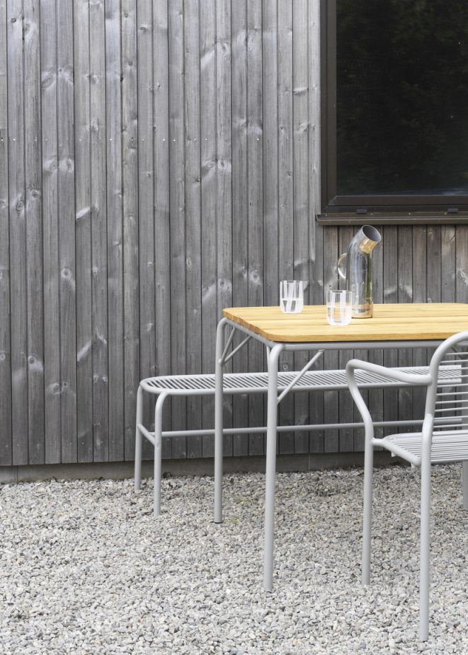Normann Copenhagen Vig Bench Bank Outdoor mit Tisch und Gläsern vor grauer Holzwand.