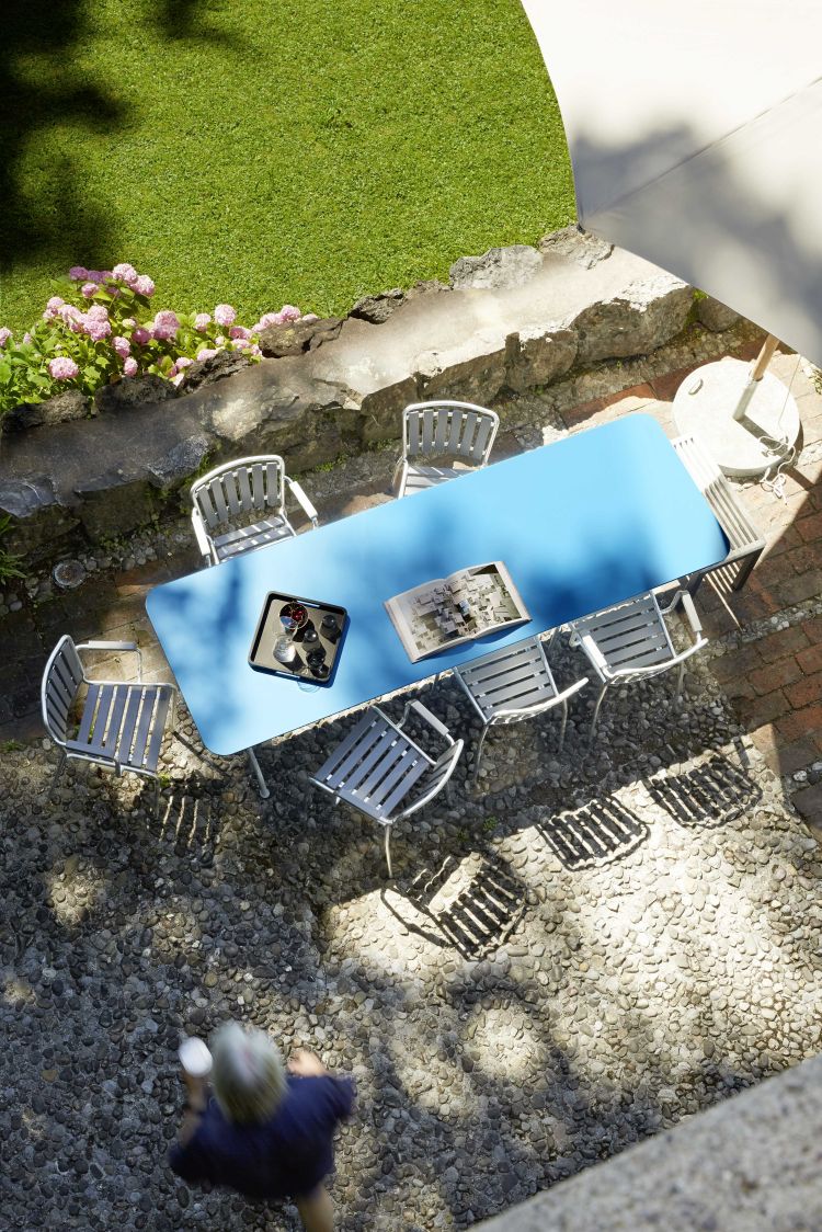 Blauer Gartentisch mit sechs grauen Stühlen, Tablett mit Getränken und Buch im Außenbereich.