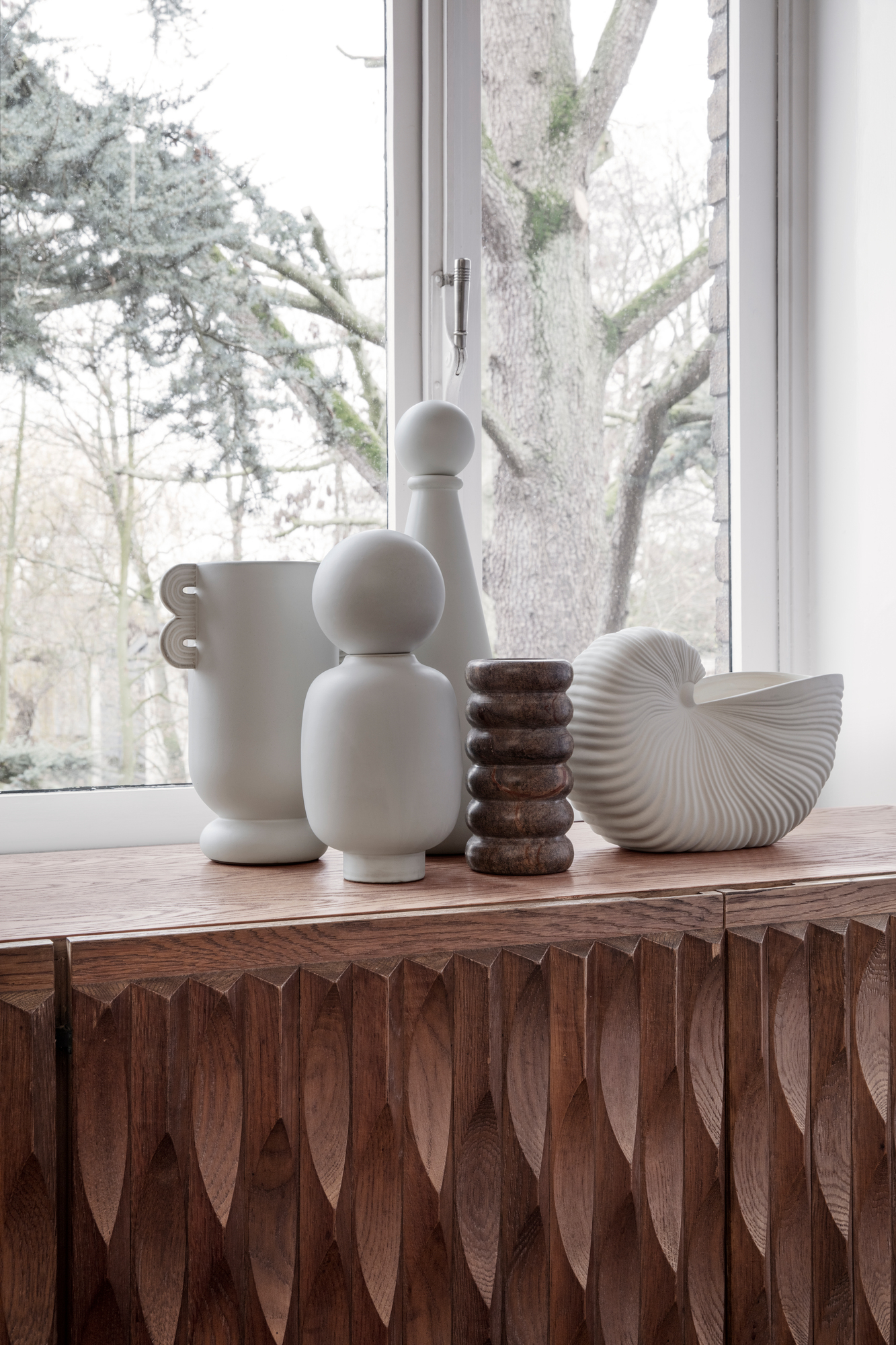 Verschiedene Vasen, darunter eine Shell Pot Vase, dekorieren ein Sideboard vor einem Fenster.