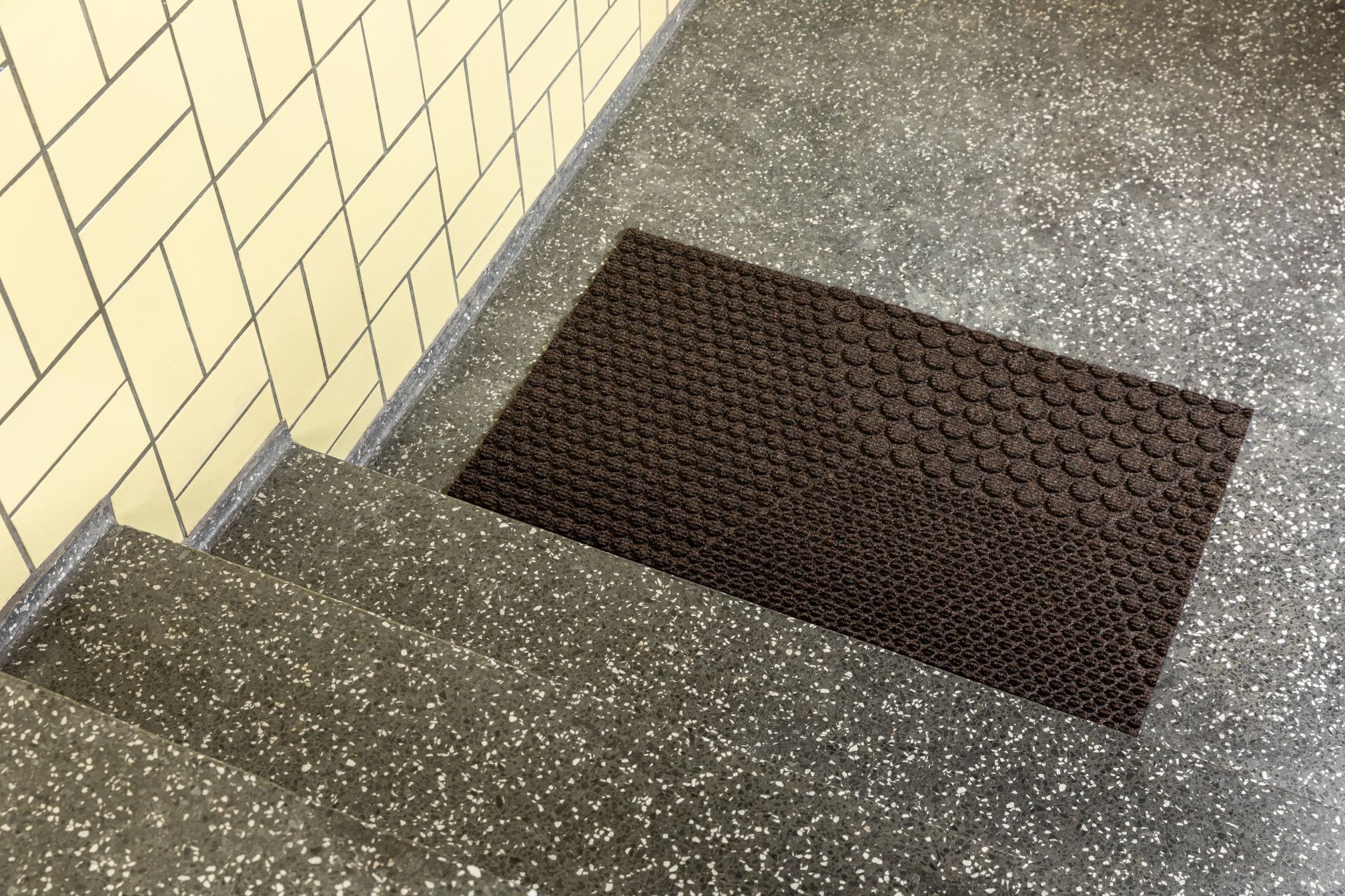 Braune Hex Fußmatte Soil von Heymat auf einer Treppe mit Terrazzo-Boden und gelben Fliesen.