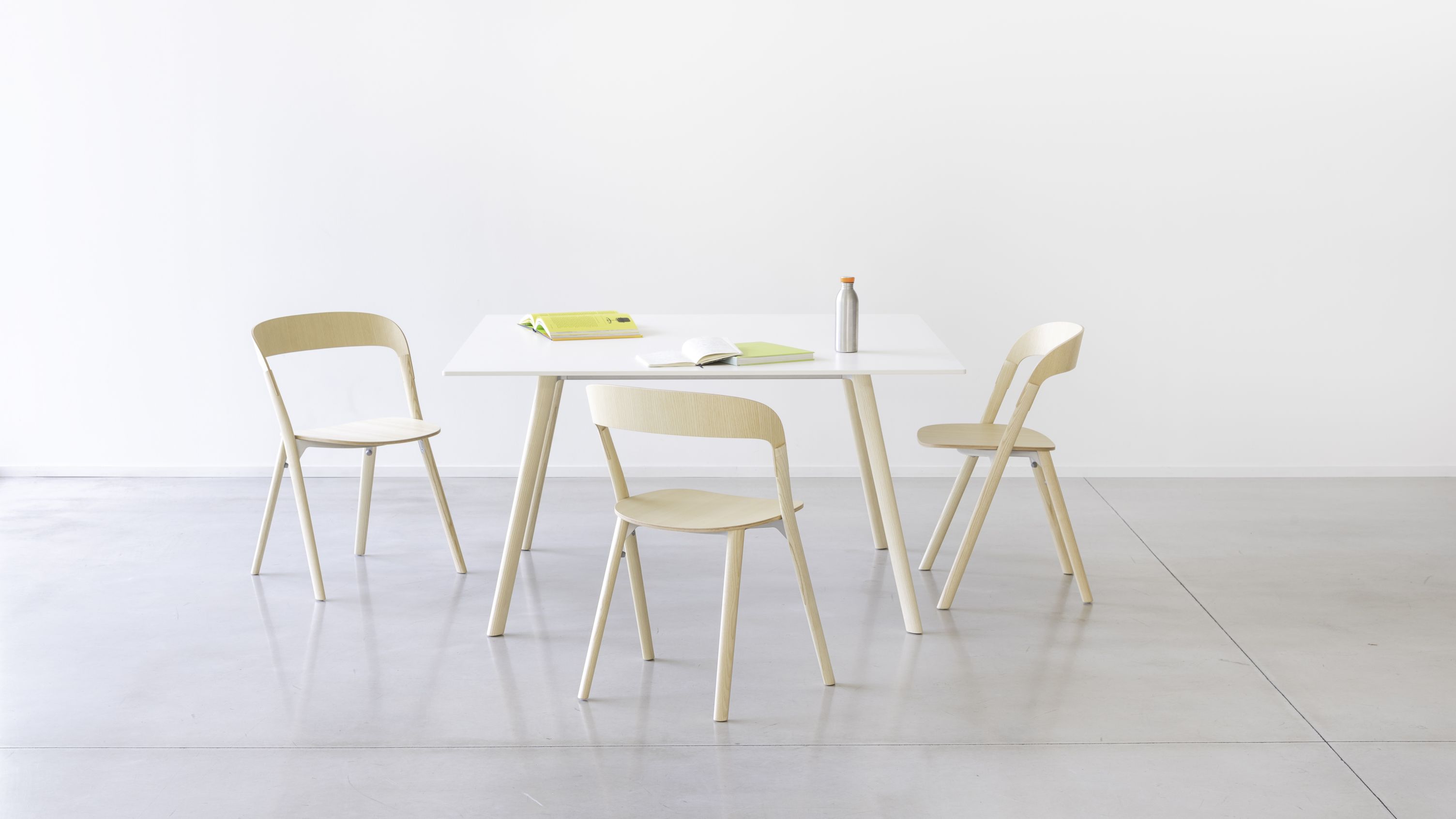 Drei helle Holzstühle vom Typ Pila Stuhl um einen weißen Tisch im minimalistischen Design.