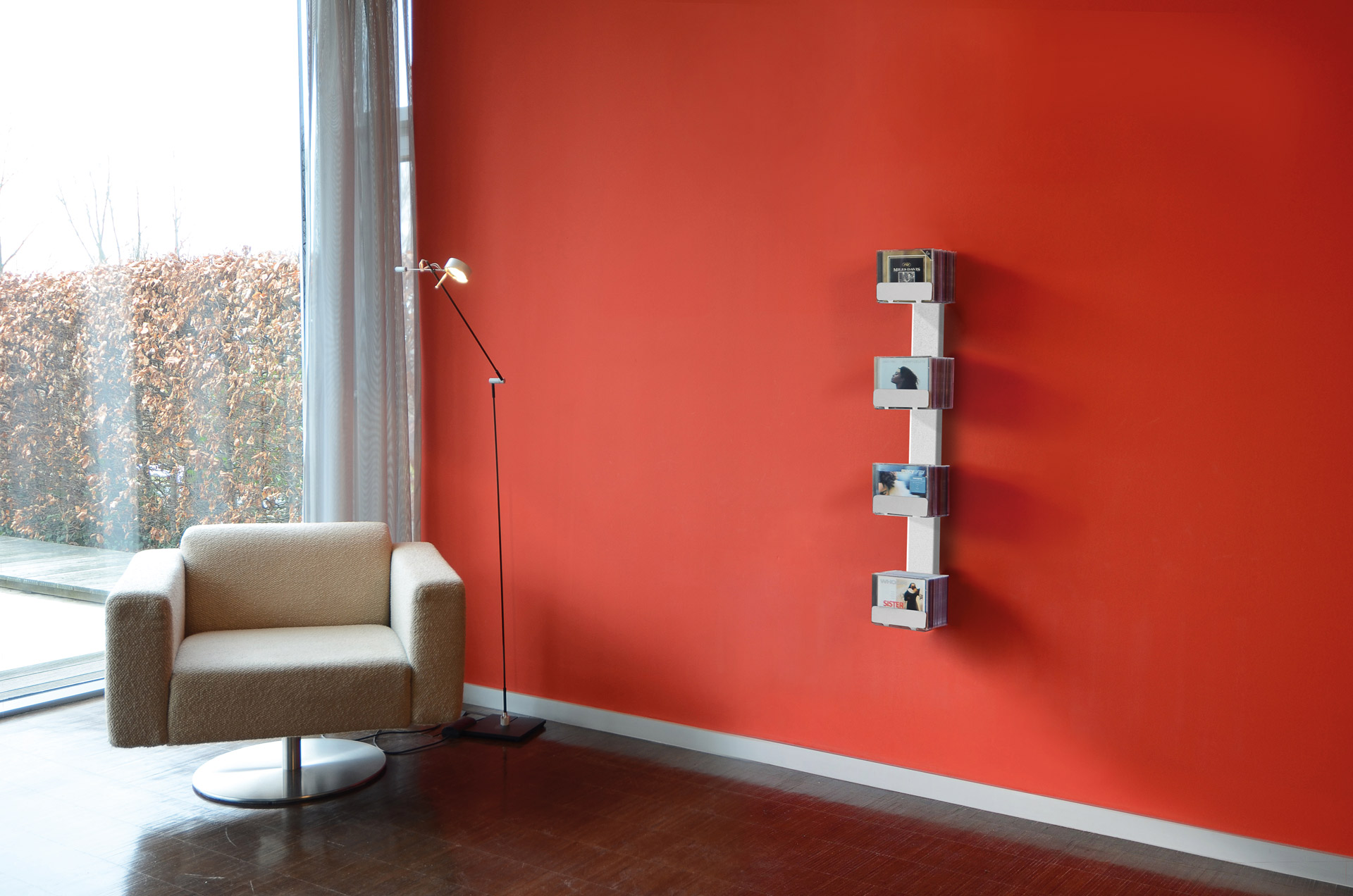 CD-Baum Wandregal: Vertikales Regal für CDs an roter Wand, neben Sessel und Lampe.
