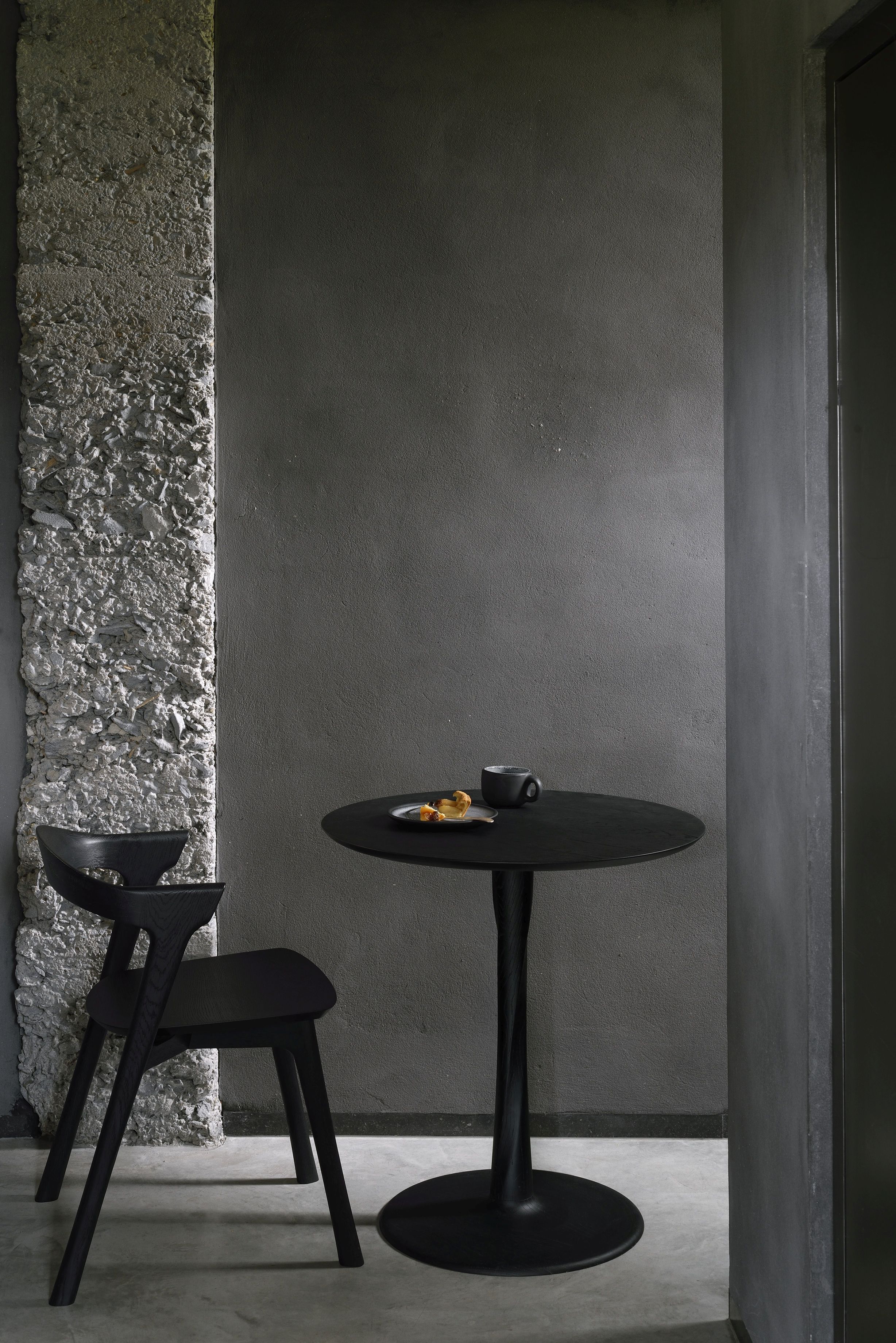 Schwarzer Ethnicraft Torsion Eiche Tisch mit Stuhl, gedeckt mit Tasse und Gebäck vor grauer Wand.
