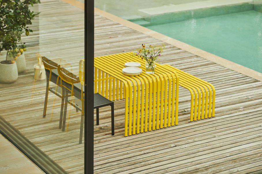 Gelber Jan Kurtz Campus Tisch Outdoor auf Holzterrasse mit Geschirr und Blumenvase.