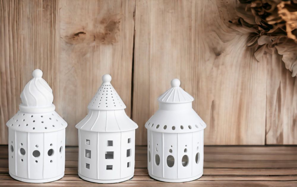 Drei weiße, dekorative Windlichter aus Keramik auf einem Holztisch vor einer Holzwand.