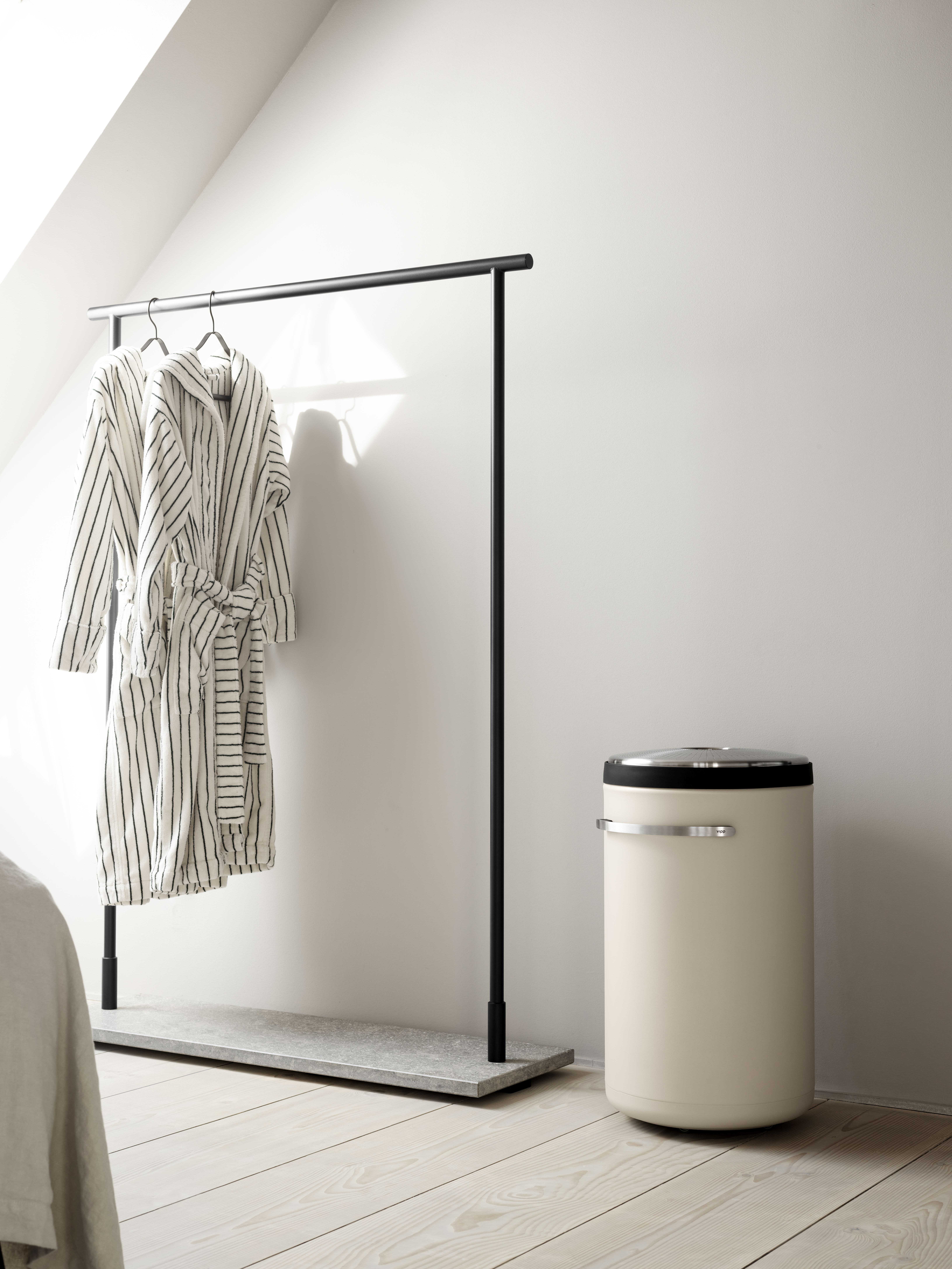 Beige Vipp Wäschekorb im minimalistischen Badezimmer neben Kleiderständer mit Bademänteln auf hellem Holzboden.