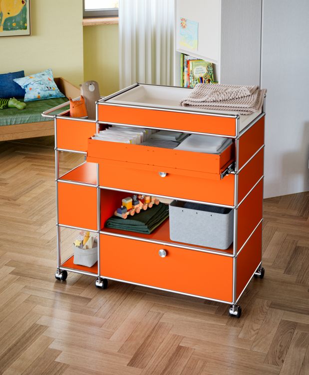Orangefarbener USM Haller Wickeltisch mit Schubladen, Ablageflächen und Rollen im Kinderzimmer.