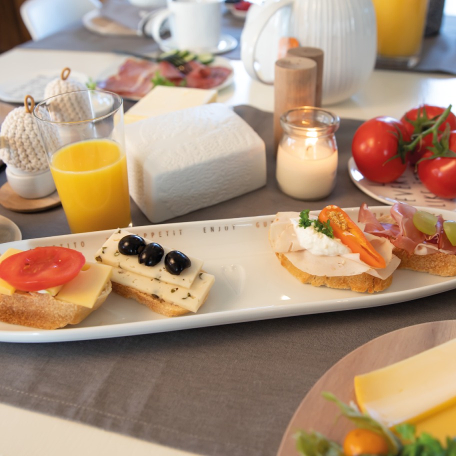 Guten Appetit Servierplatte von Räder mit belegten Broten, Käse und Tomaten auf gedecktem Frühstückstisch.