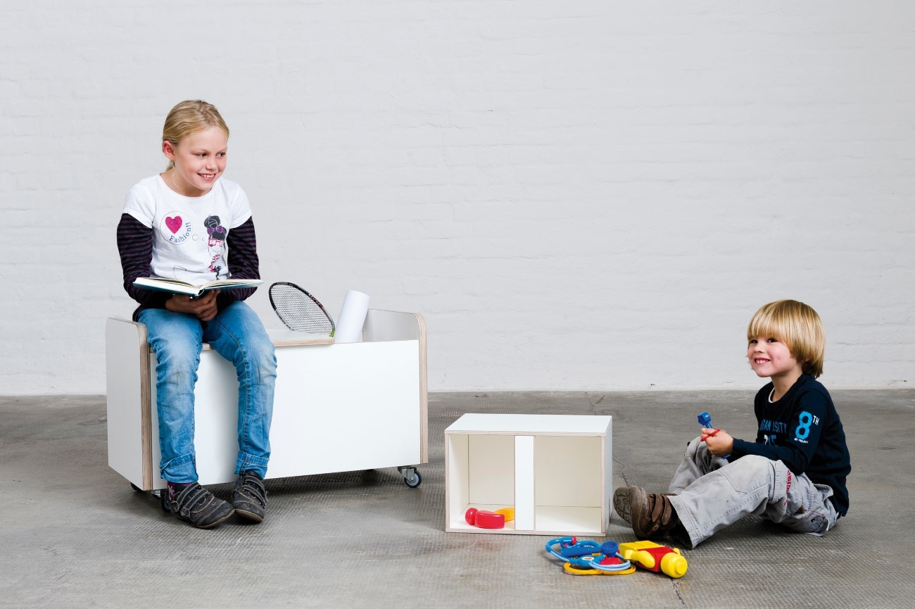 Mädchen liest auf Growing Table Rollbox Truhe, Junge spielt mit Spielzeug neben kleiner Truhe.