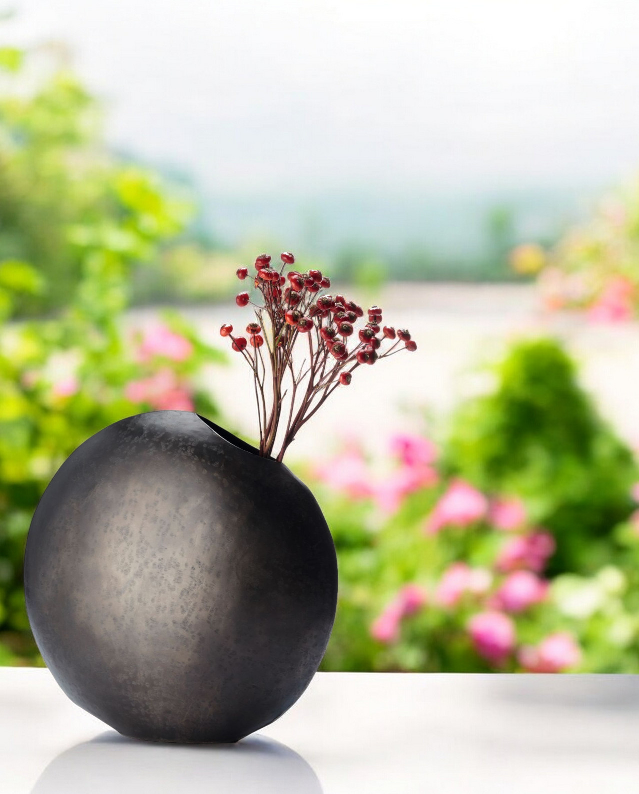Monrovia Vase Philippi: Runde, dunkle Design-Vase mit roten Beeren-Zweigen vor verschwommenem Gartenhintergrund.