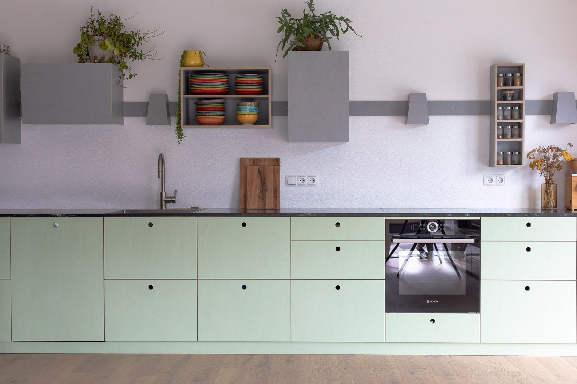 Helle Küche mit mintgrünen Unterschränken, schwarzer Arbeitsplatte und Bosch Backofen. Moderne Kücheneinrichtung mit Regalen und Dekoration.