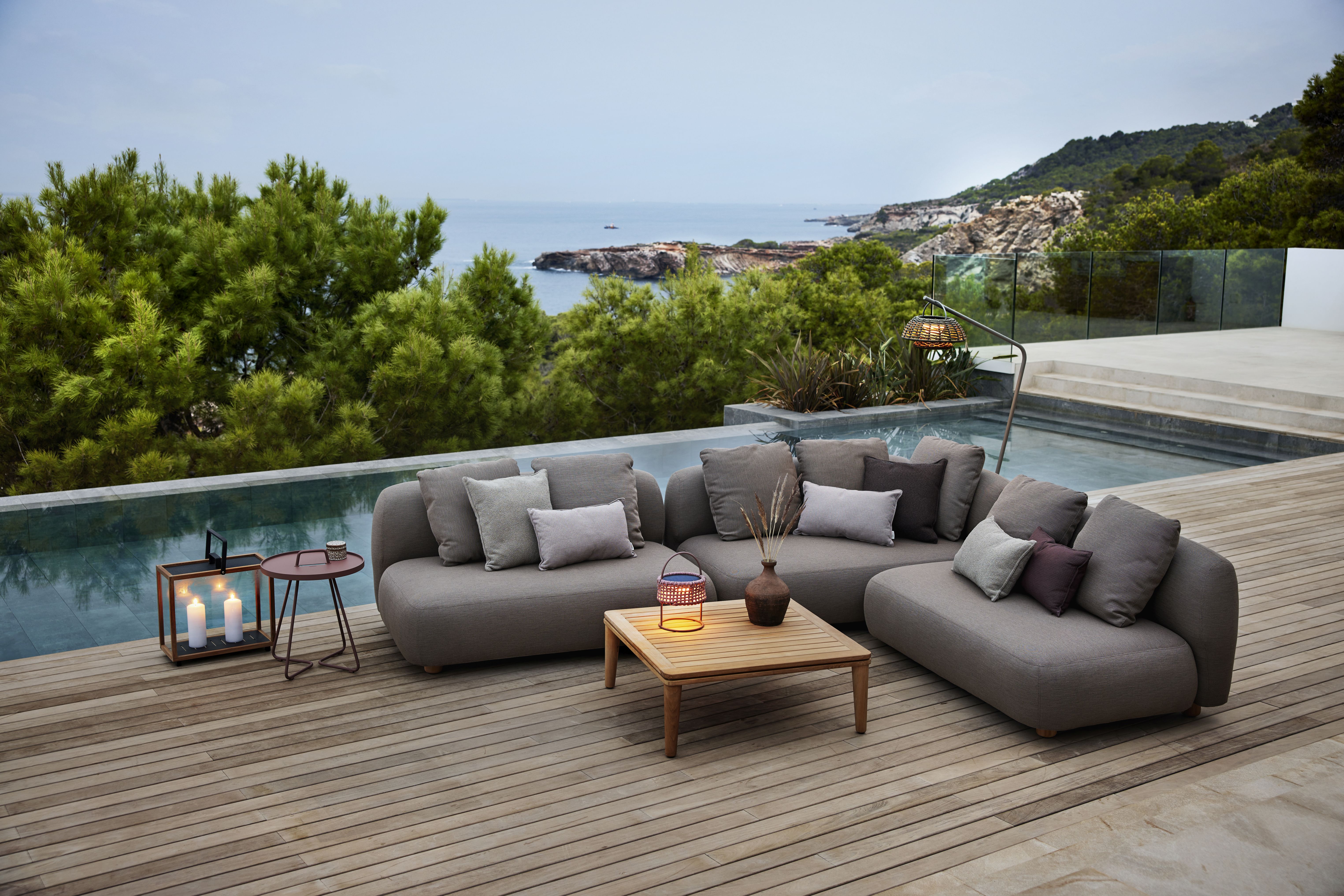 Cane-Line Capture Outdoor Couchtisch aus Teakholz neben grauer Lounge-Garnitur mit Kissen auf Holzterrasse.