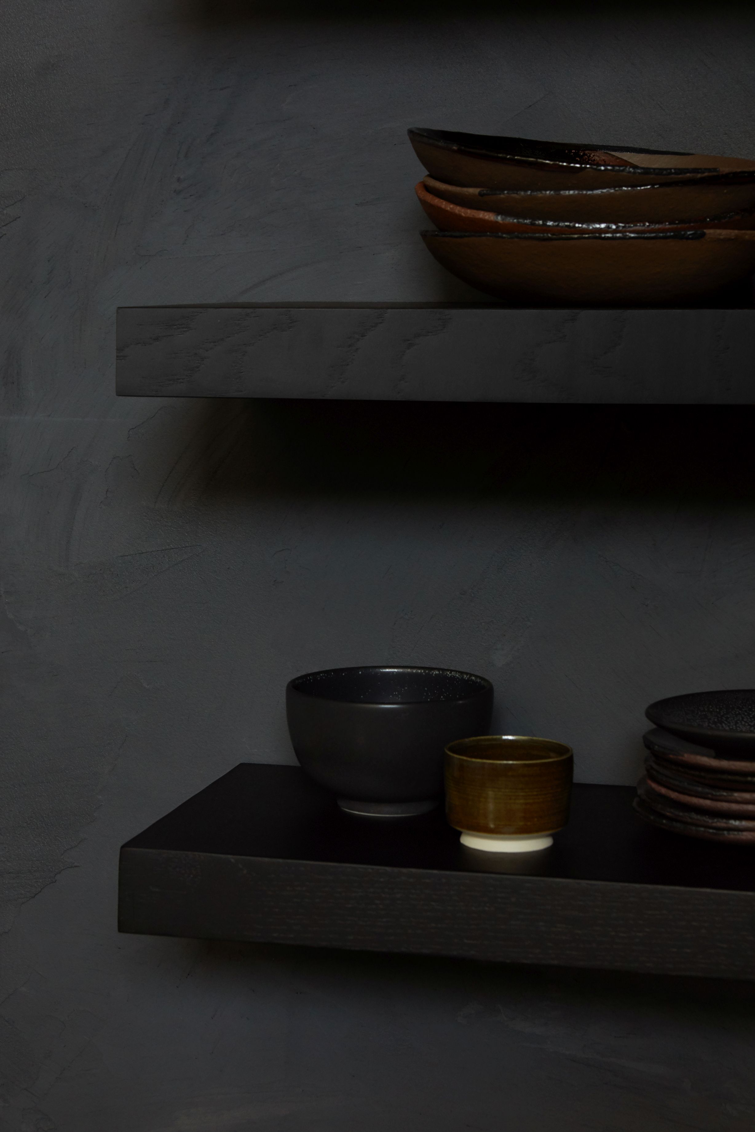 Schwarzes Ethnicraft Wandregal aus Eiche mit Geschirr, Schalen und Tassen auf zwei Ebenen vor dunkler Wand.