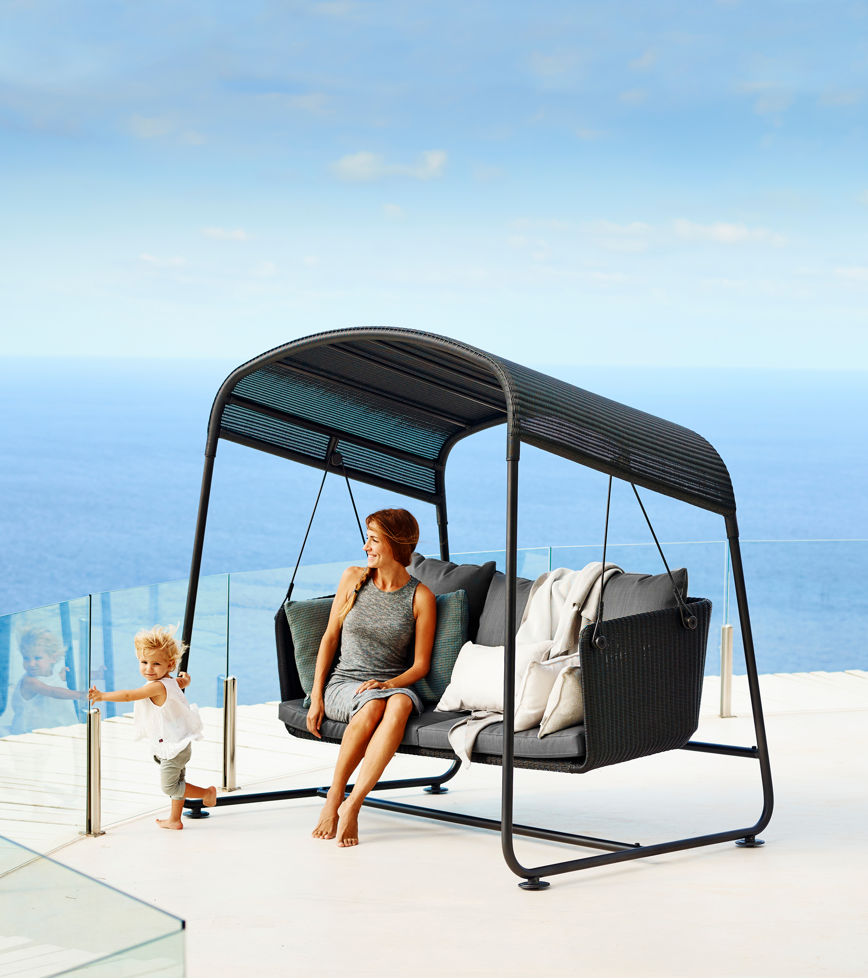 Frau entspannt auf Cane-Line Gartenschaukel "Cave" mit Kissen vor Meerblick.