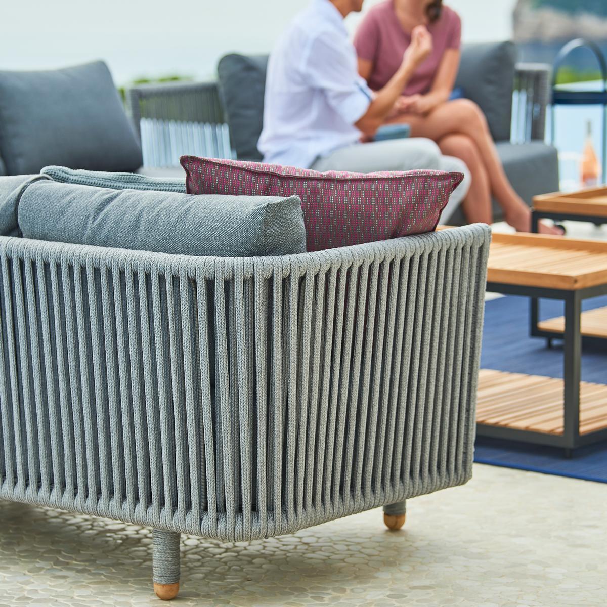 Graues Cane-Line Moments Outdoor Sofa mit Kissen und zwei Personen im Hintergrund.